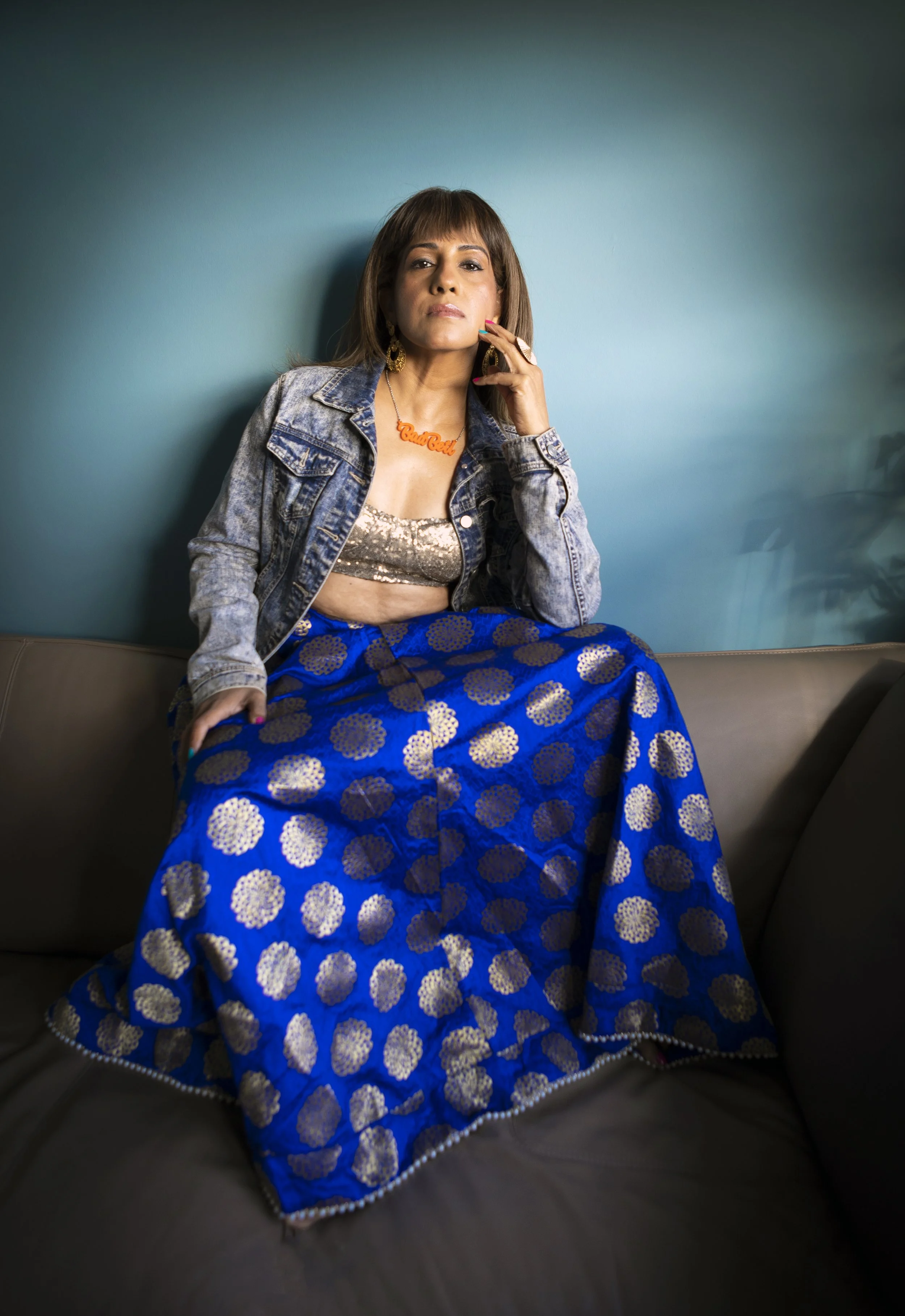 Woman in a denim jacket and a blue traditional dress with gold patterns, sitting on a dark-colored couch against a teal wall, accessorized with gold earrings, a necklace with orange lettering, and rings.