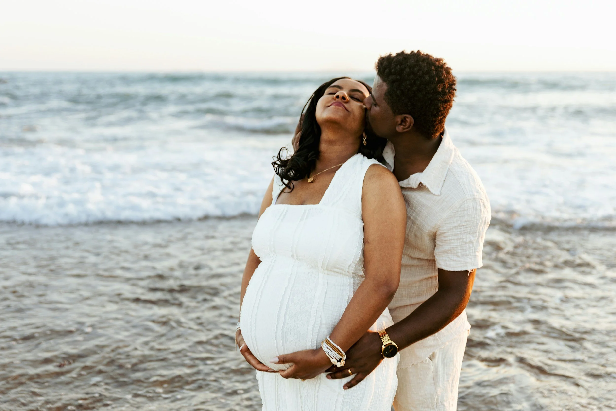 A pregnant woman in a white dress being embraced from behind by a man on the beach, both smiling with the ocean in the background.