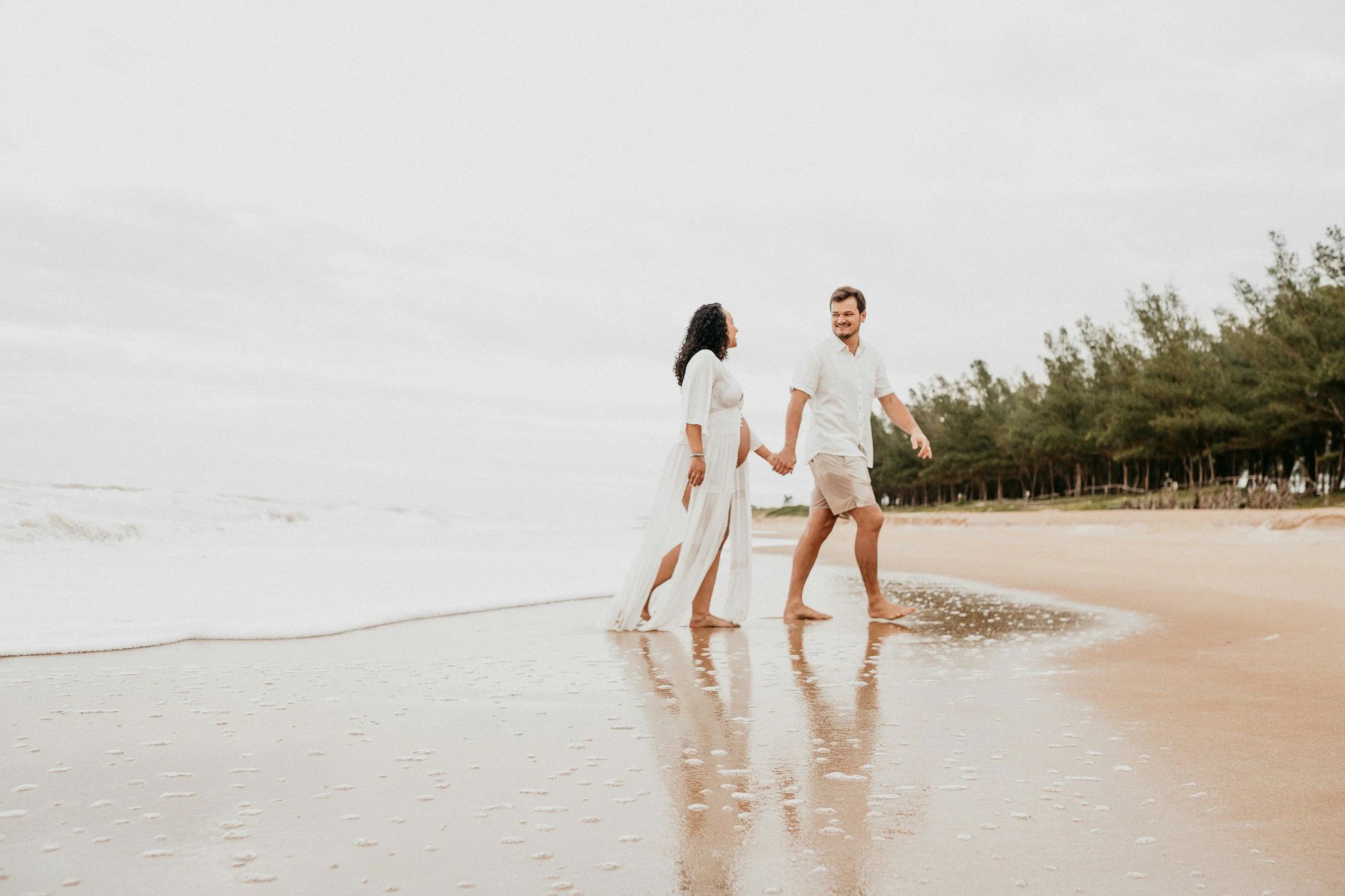 A couple holding hands and walking along the shoreline at a beach with trees in the background, overcast sky.