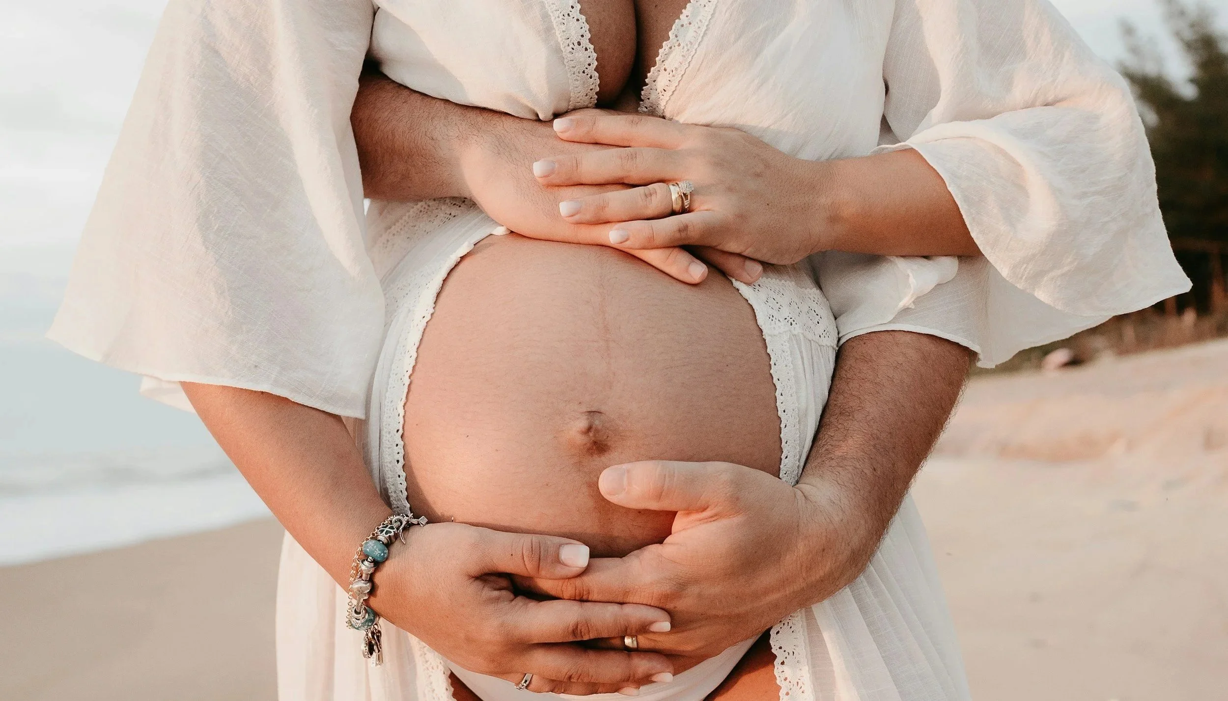 A pregnant woman is being embraced from behind by a man, both with their hands on her belly. They are outdoors, possibly on a beach. The woman is wearing a white, lacy, loose dress, and the man is shirtless. Some jewelry is visible, including rings and a bracelet.