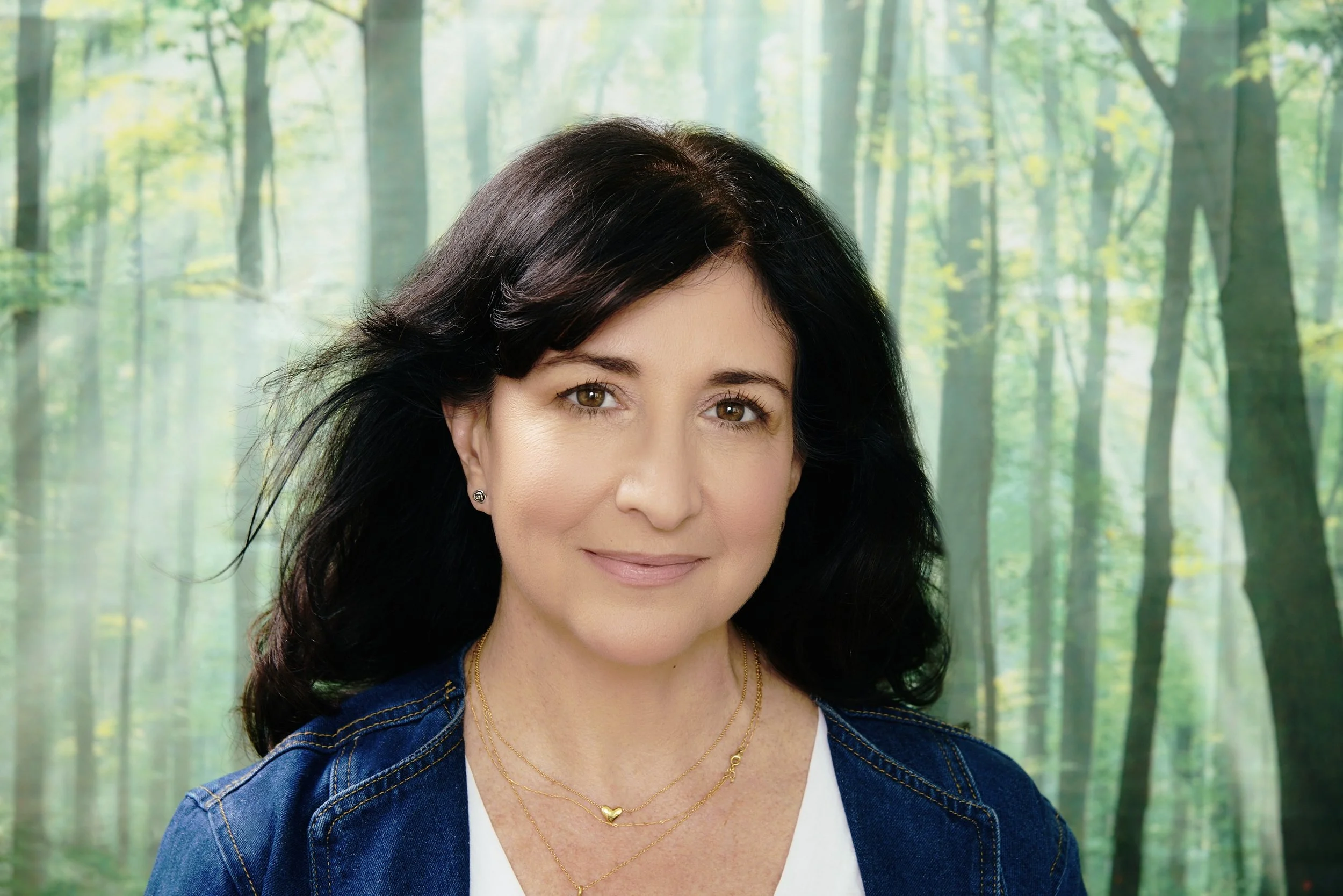 Portrait of a woman with dark hair, wearing a denim jacket, white top, and gold jewelry, standing outdoors with a blurred green forest background.