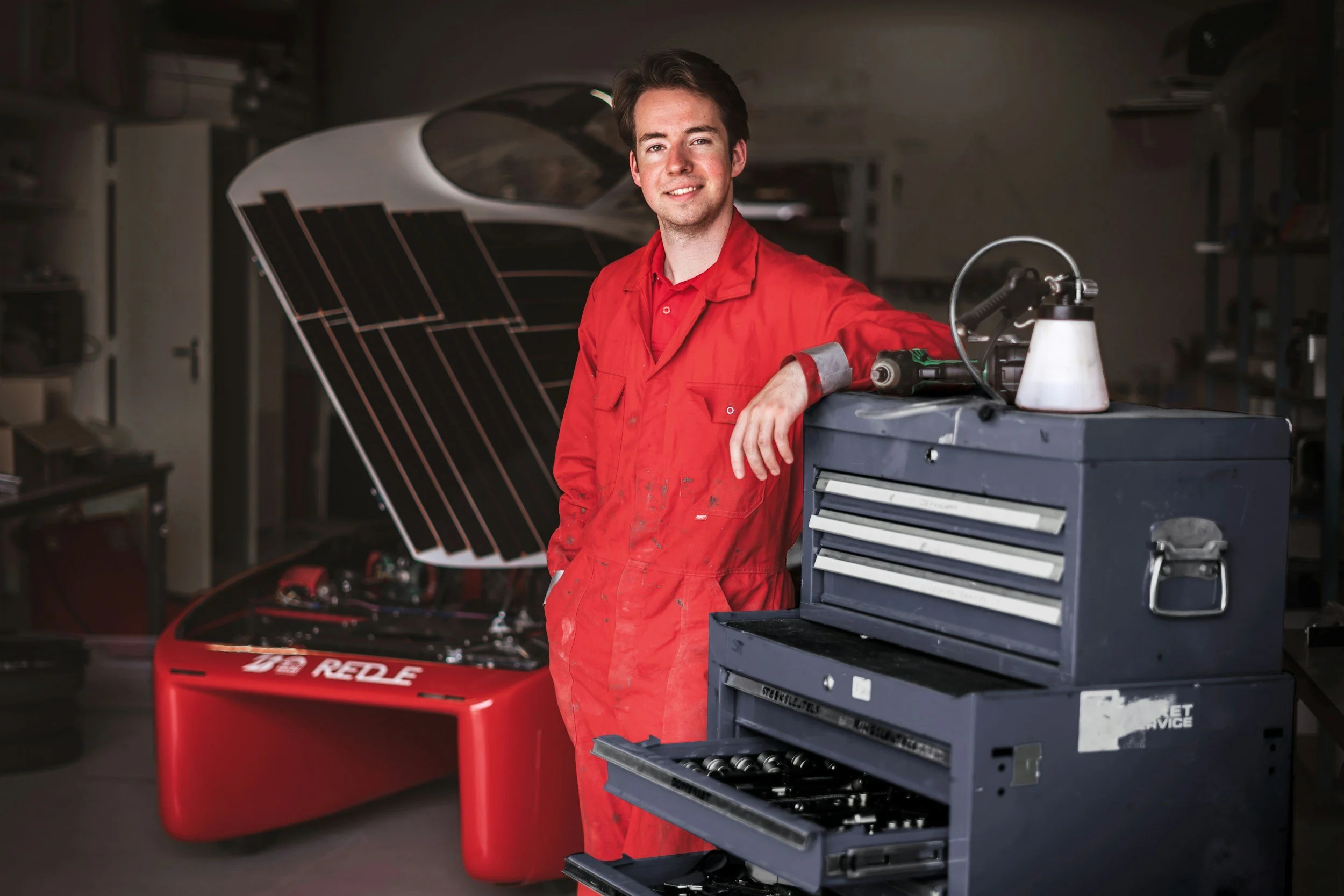 Young man in red mechanic coveralls standing in a garage with a car in the background, smiling and leaning on a blue tool chest.
