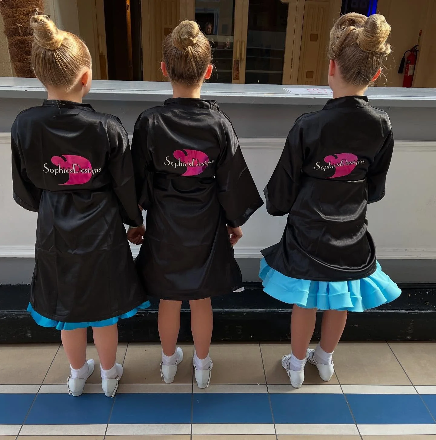 Three young girls standing with their backs facing the camera, wearing matching black jackets with a pink heart logo and blue skirts, in a public indoor setting.