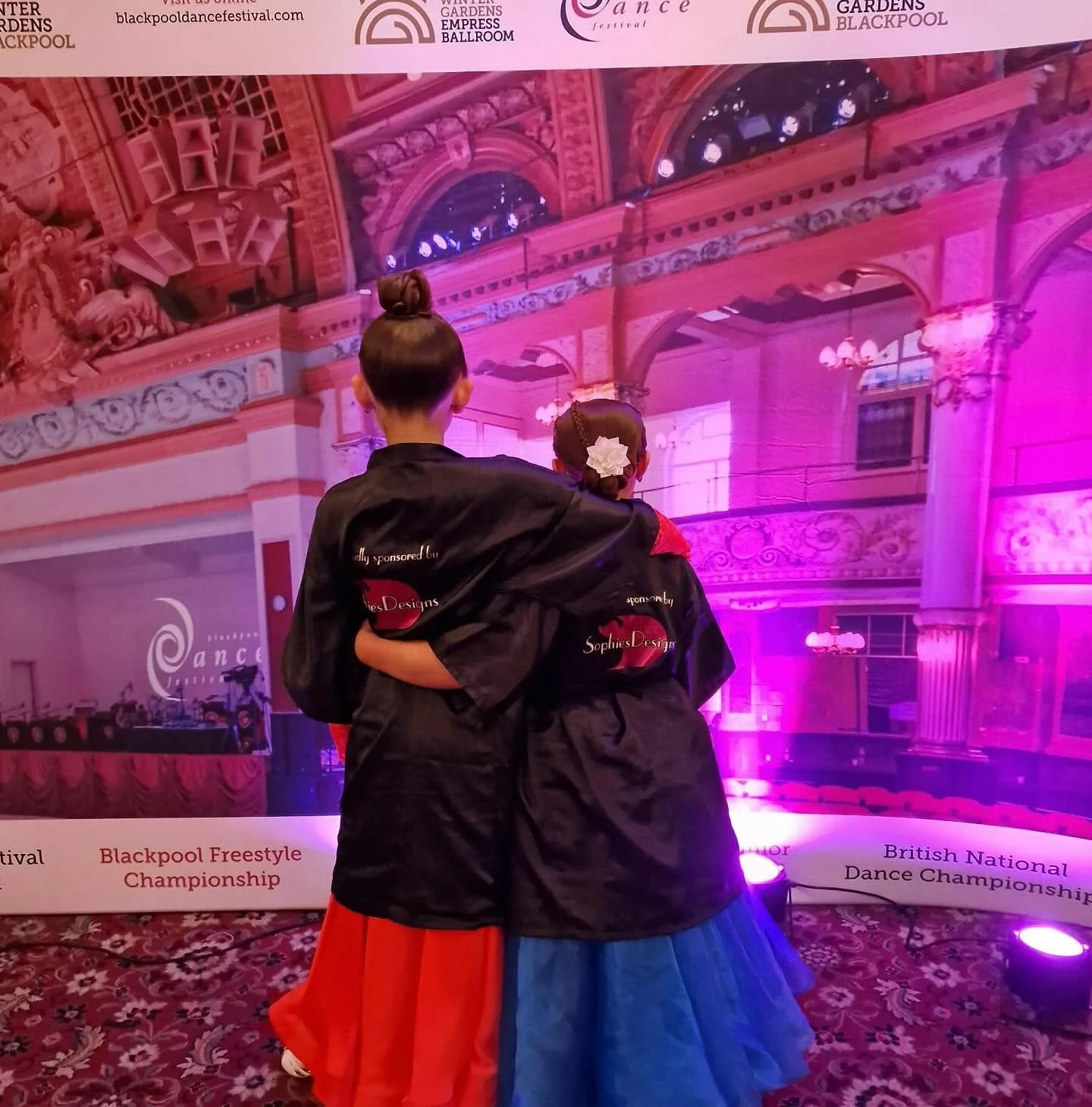 Two young dancers in black jackets and colorful skirts, with their arms around each other, stand in front of a pink-lit stage with ornate architecture, at the Blackpool Freestyle Championship, a British National Dance Championship.