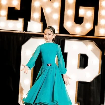 Young girl in a teal dress smiling on stage with large illuminated letters in the background.