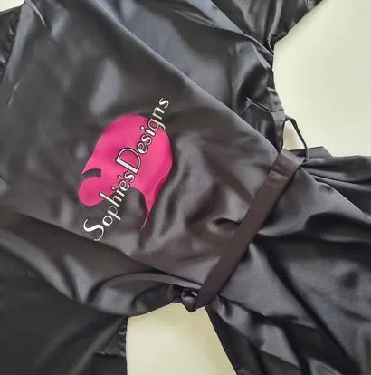 Black satin garment with embroidered pink logo of Marie Antoinette, with the text 'Sophie’s Designs'.