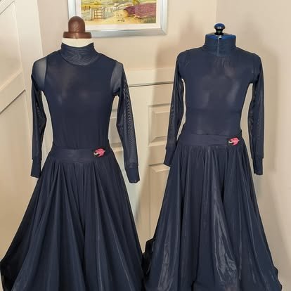 Two mannequin dress forms wearing navy blue long-sleeve dresses with turtleneck collars and full skirts, displayed in a room with a picture frame on the wall.