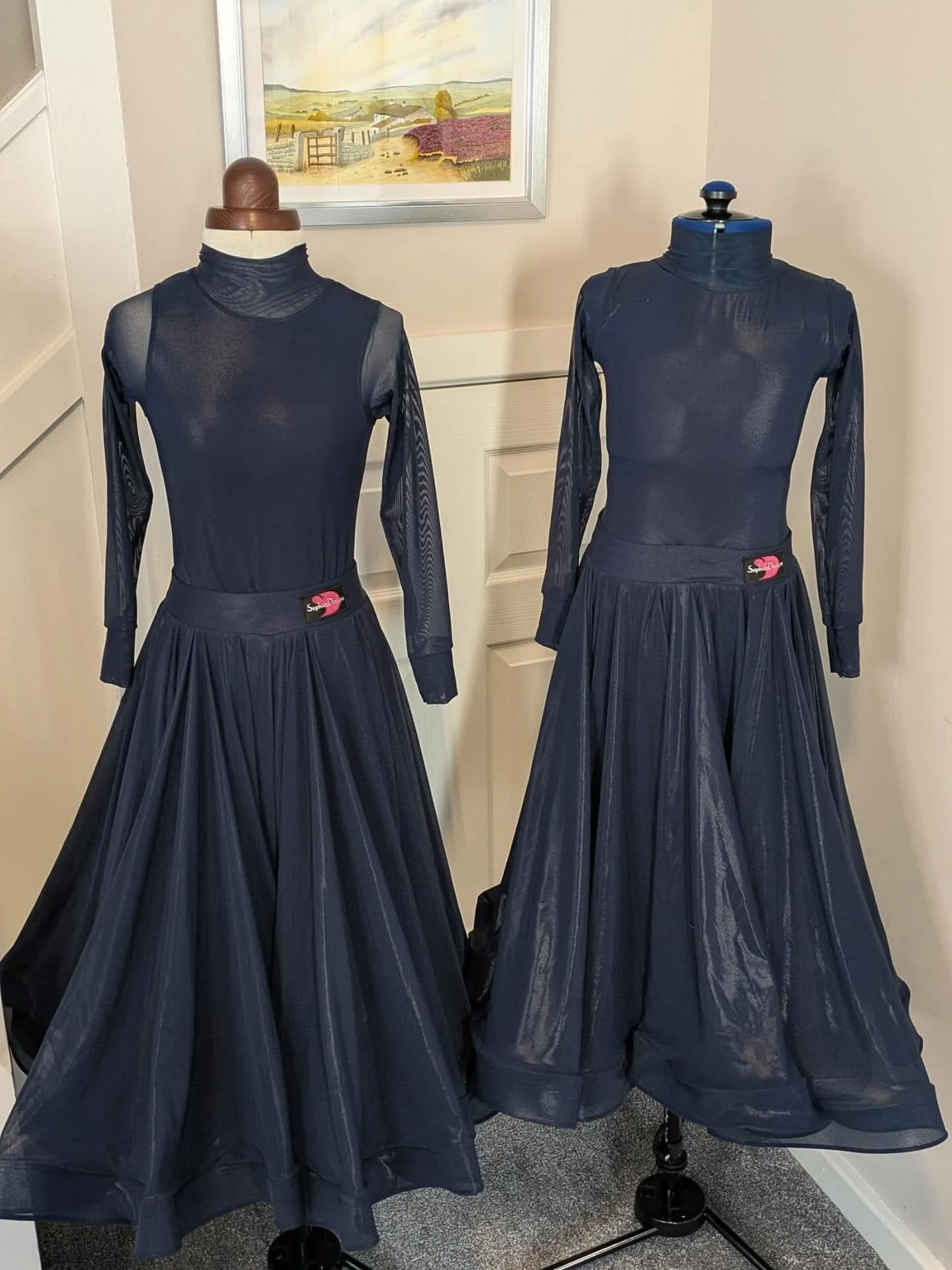 Two mannequins dressed in matching black long-sleeve tops with turtlenecks and black pleated skirts on a display stand, in front of a framed landscape painting.