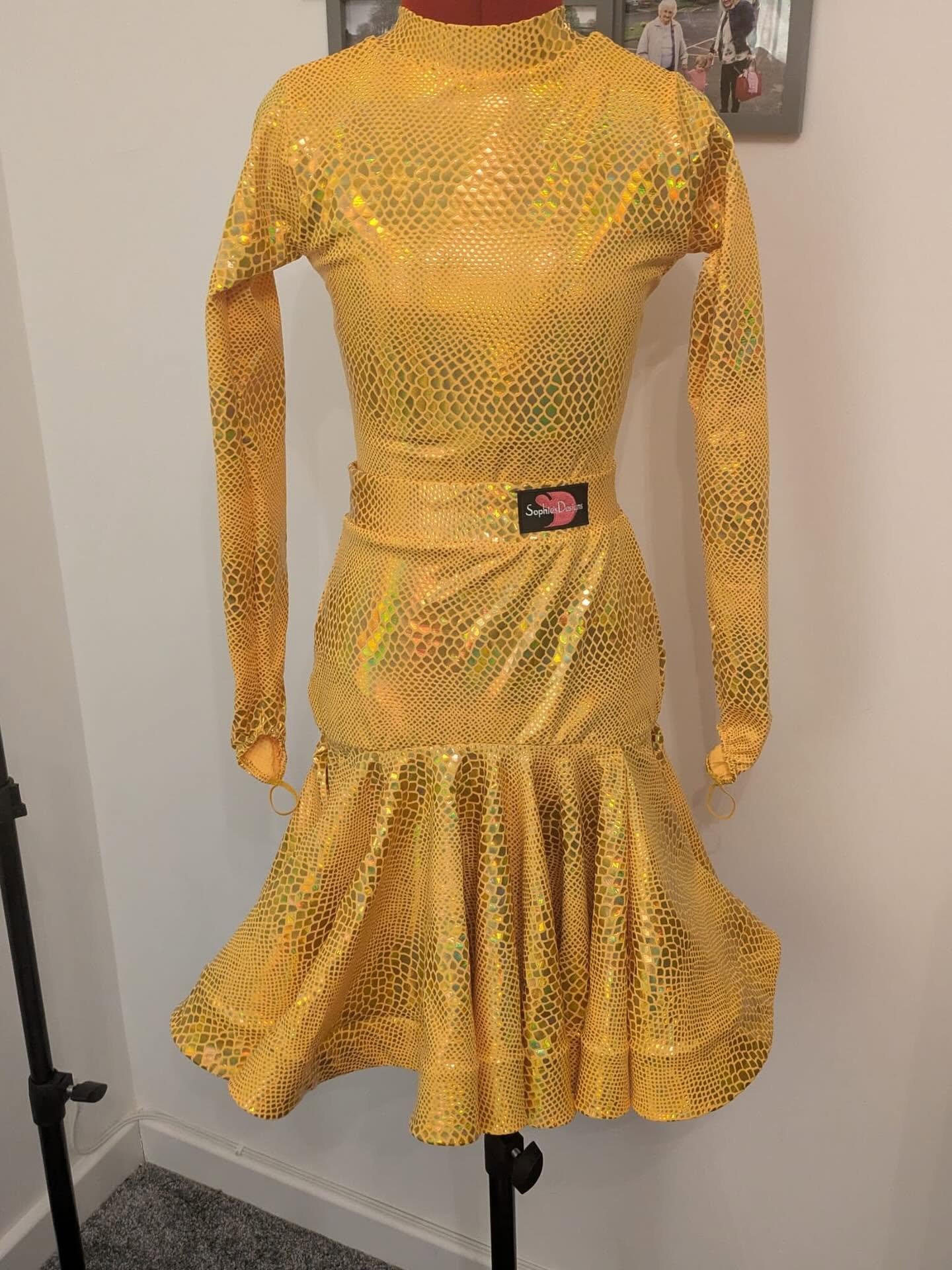 A mannequin wearing a shiny yellow, crocodile-patterned dress with long sleeves, a fitted waist, and a ruffled hem, displayed on a black stand against a light-colored wall with a framed photo in the background.