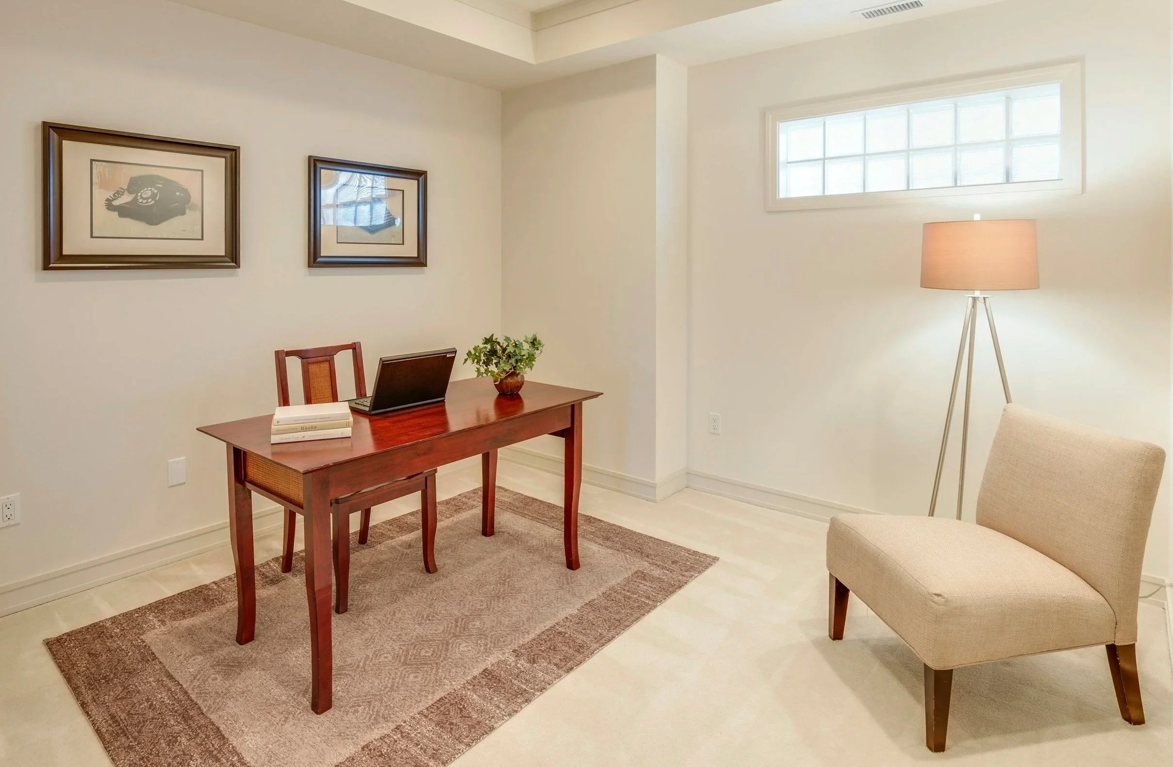 A small, minimalist home office with a wooden desk holding a laptop, books, and a potted plant, with two framed art pieces on the white wall, a beige chair, a floor lamp, a window with frosted glass, and a beige armchair.