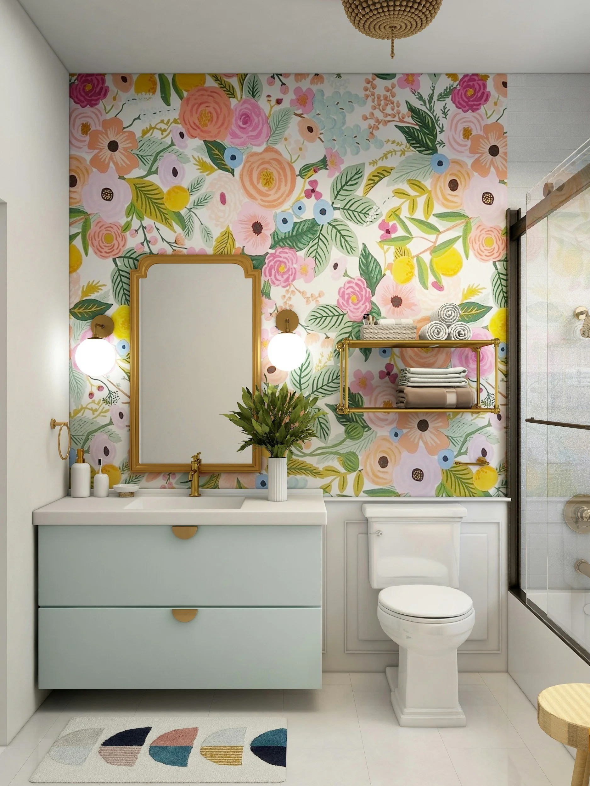 A modern bathroom with floral wallpaper featuring pink, peach, yellow, and blue flowers with green leaves. There is a white vanity with a built-in sink, gold hardware, and a large mirror above. A vase with green plants is on the vanity. To the right, there is a toilet and a glass shower enclosure. Gold shelving holds folded towels, and a striped rug with geometric shapes is on the white floor. The ceiling light fixture is round with a decorative gold chain.