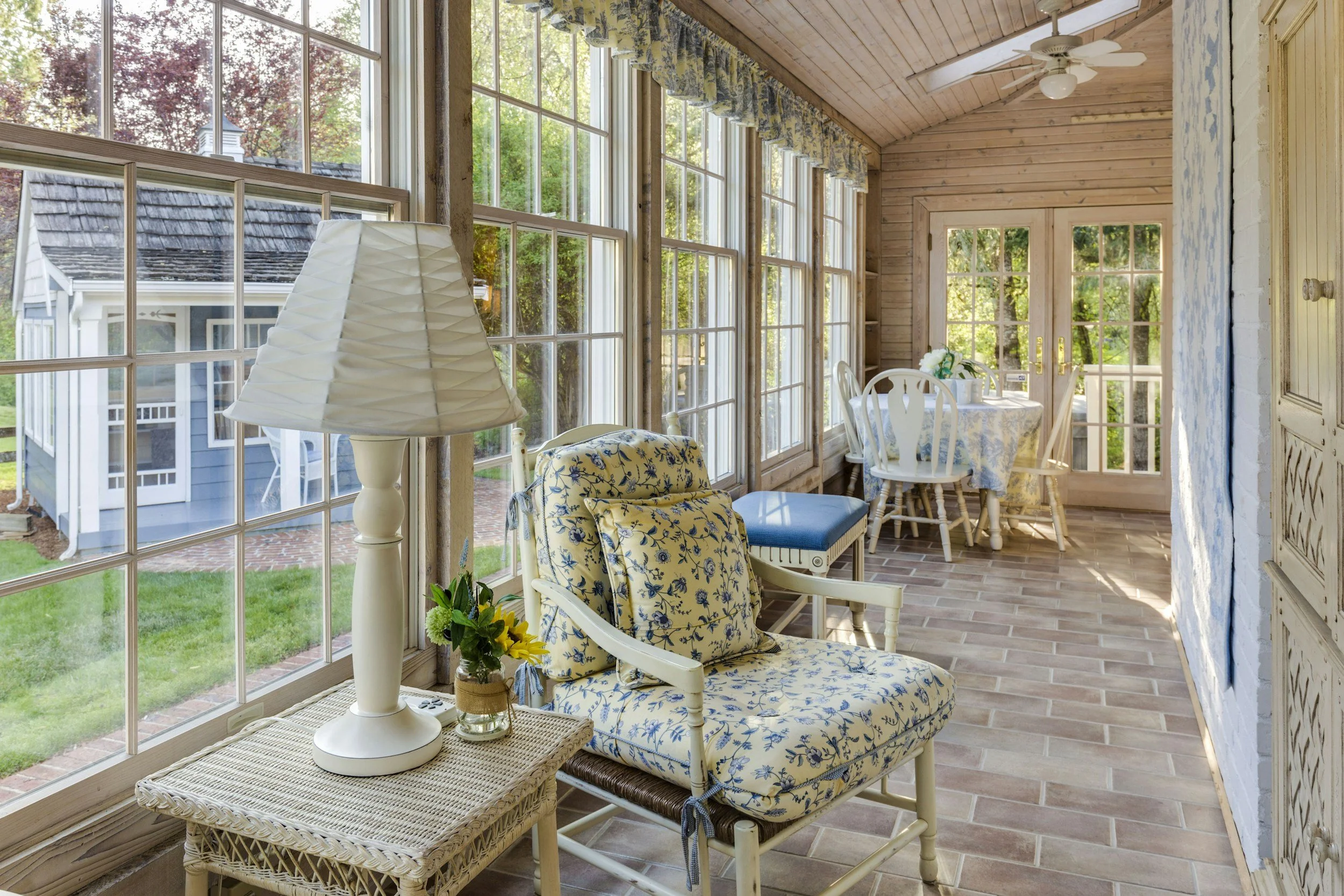 Sunroom with large windows, wooden ceiling, ceiling fan, small dining table with white chairs, floral armchair, table lamp, and decorative curtains