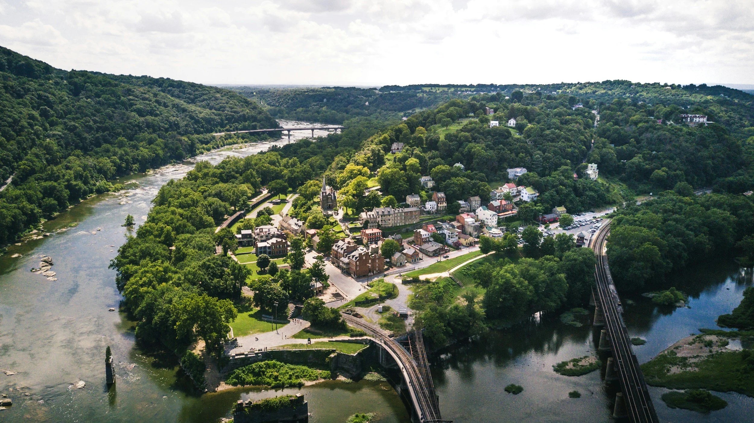 Aerial view of a river running through a lush green valley with houses and buildings along the shoreline, a bridge crossing the river, and a hillside with scattered homes and forested areas under a partly cloudy sky.