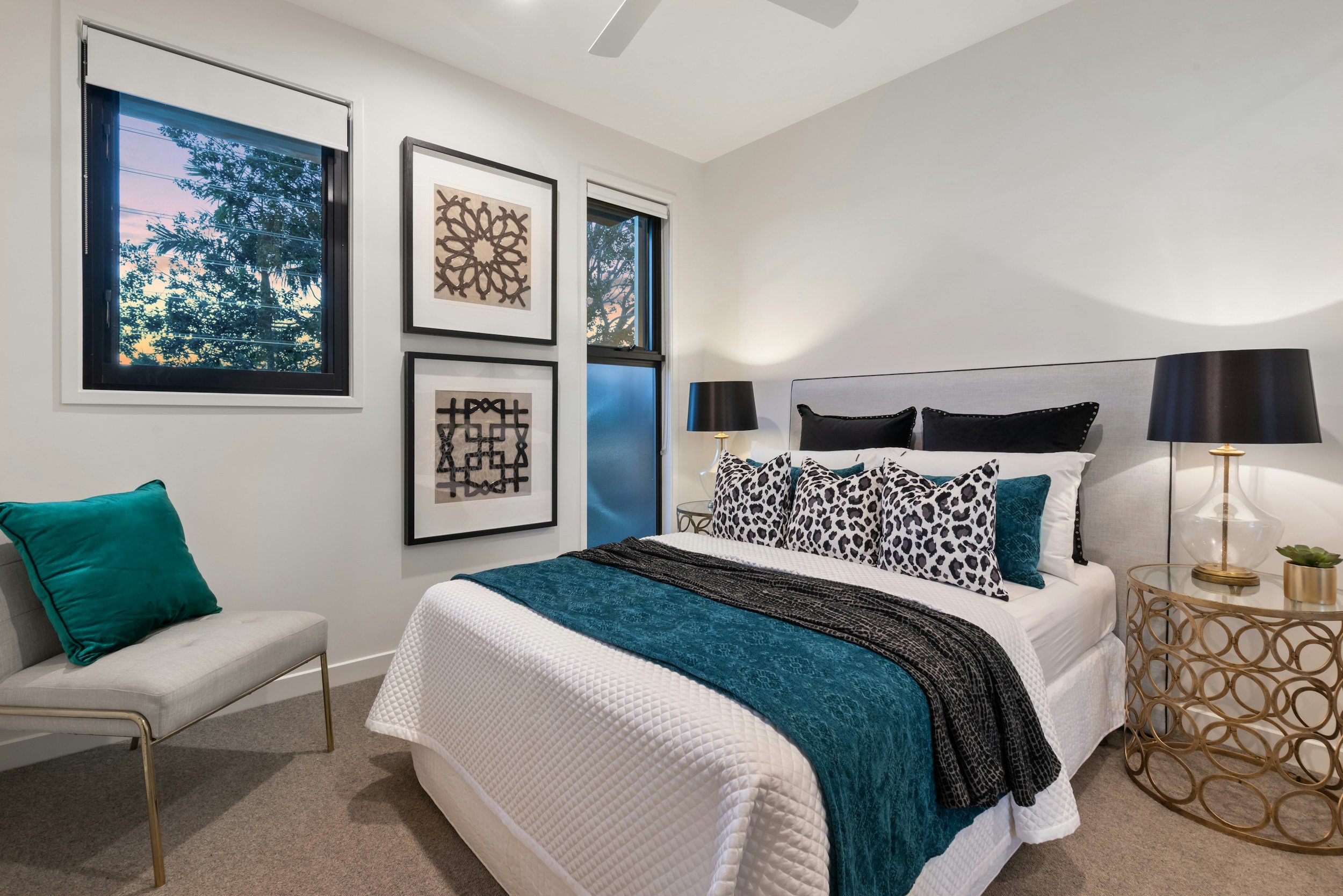 Modern bedroom with white walls, a large bed with a white quilt, teal and leopard print pillows, and black and white artwork on the wall. Two black lamps, a small round side table, and a white bench with teal and seafoam pillows are also visible.