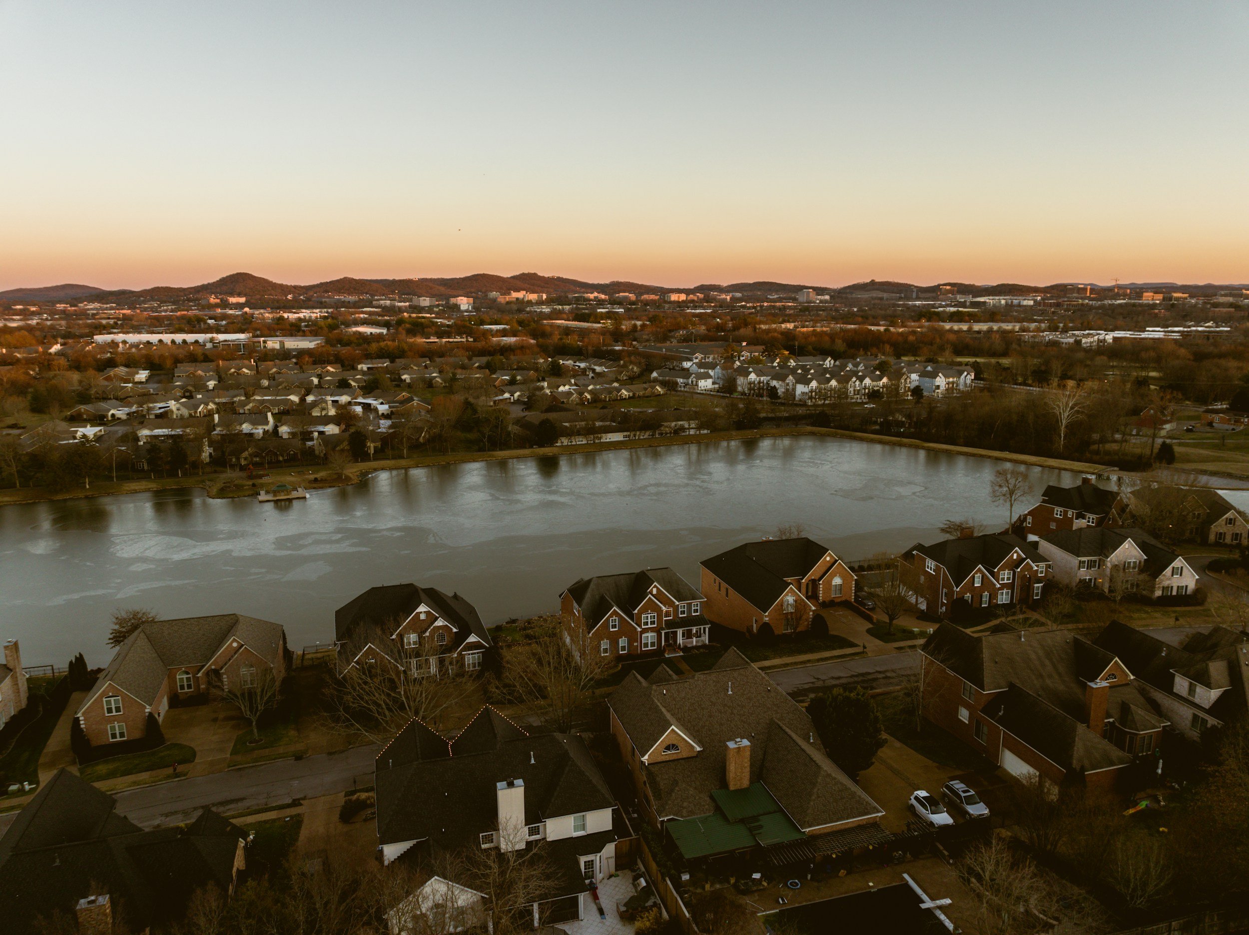 Aerial view of a suburban residential area with houses, a pond, and distant hills at sunset.