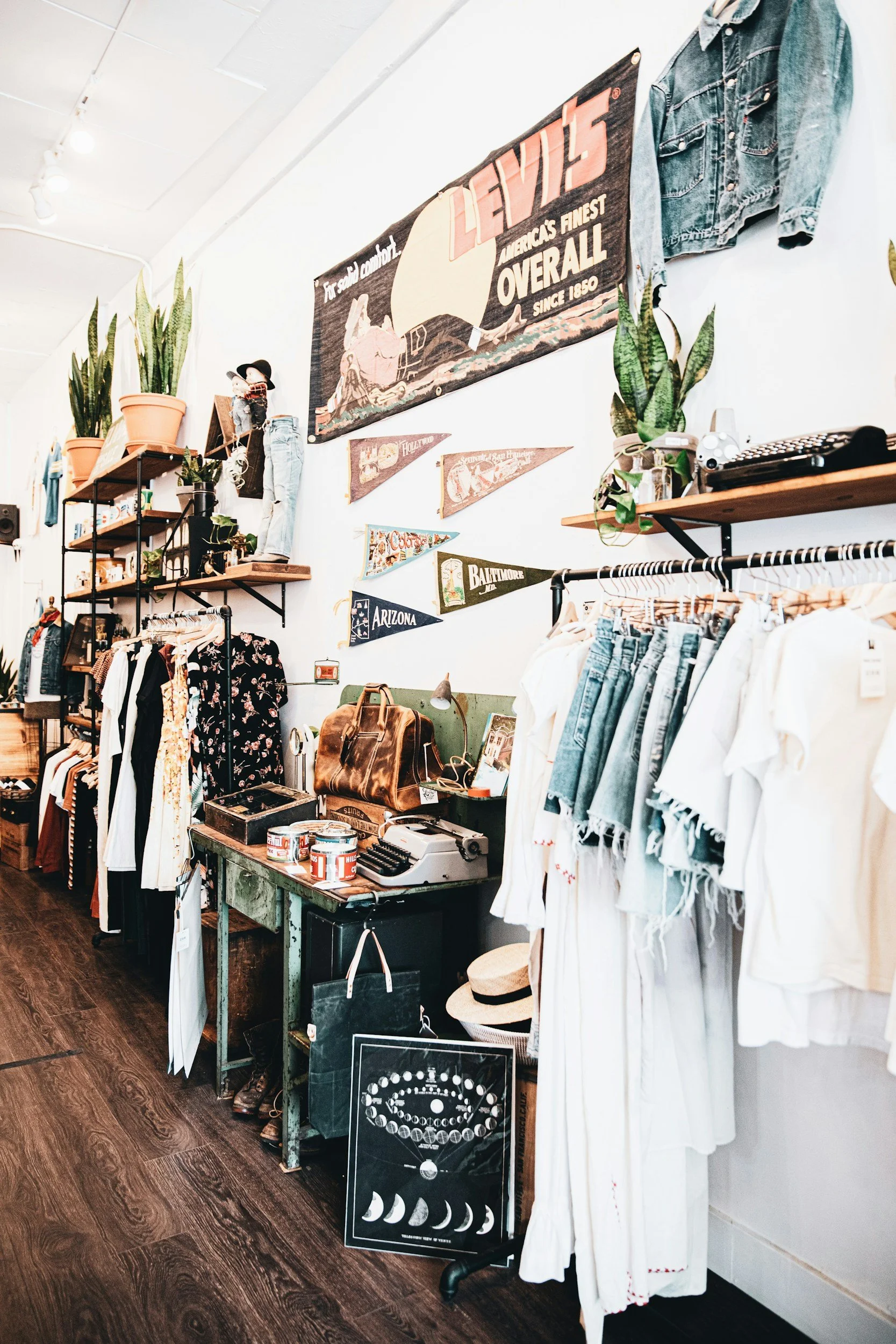 Interior of a vintage or thrift store with clothing racks, decorative banners, plants, and various accessories on display. There are shelves with books, plants, a doll, and a denim jacket hanging. On the wall, there is a large vintage Levi's banner and small decorative flags with city names.