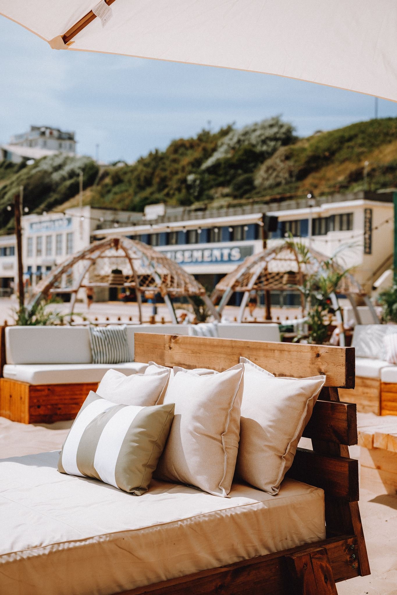 Close-up of a wooden outdoor bed with beige and white striped pillows, set on a sandy surface, with umbrellas and a hillside with buildings in the background.