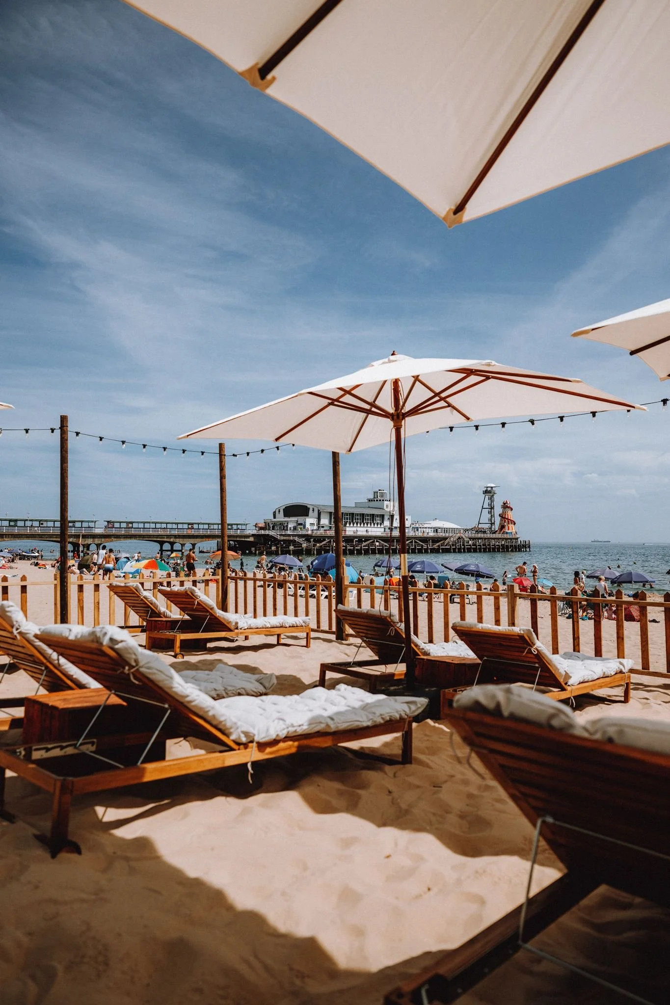 Beach scene with lounge chairs and umbrellas under blue sky, pier with building and lighthouse in the background, people relaxing and walking on the sand.