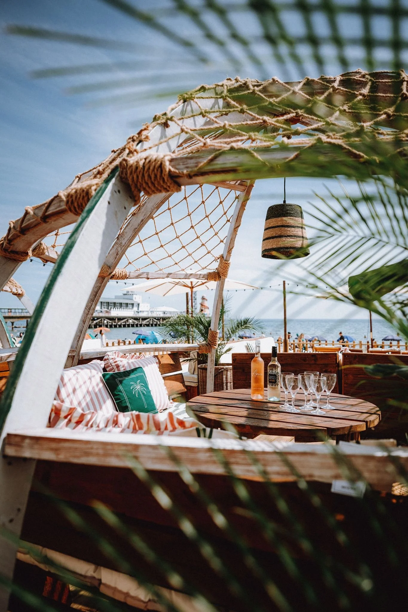 Beachside outdoor seating area with a wooden table, glass bottles, and glasses, shaded by a striped ceiling, overlooking the ocean with a pier and umbrellas in the background, surrounded by tropical plants.
