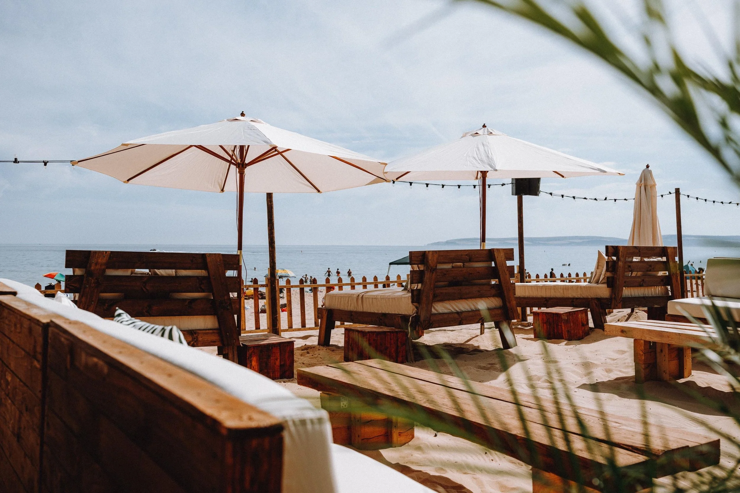 Beach scene with large white umbrellas, wooden benches, and people swimming in the ocean.