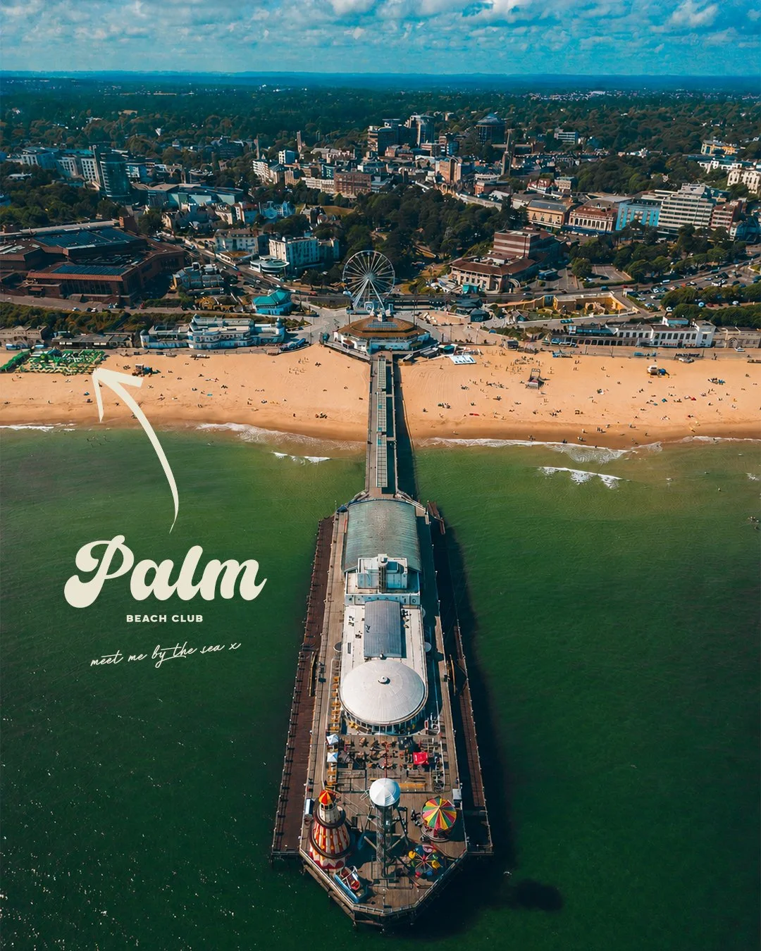 Aerial view of the Palm Beach Boardwalk and beach with a pier extending into the ocean, featuring a small amusement park with colorful rides, and a city skyline in the background.