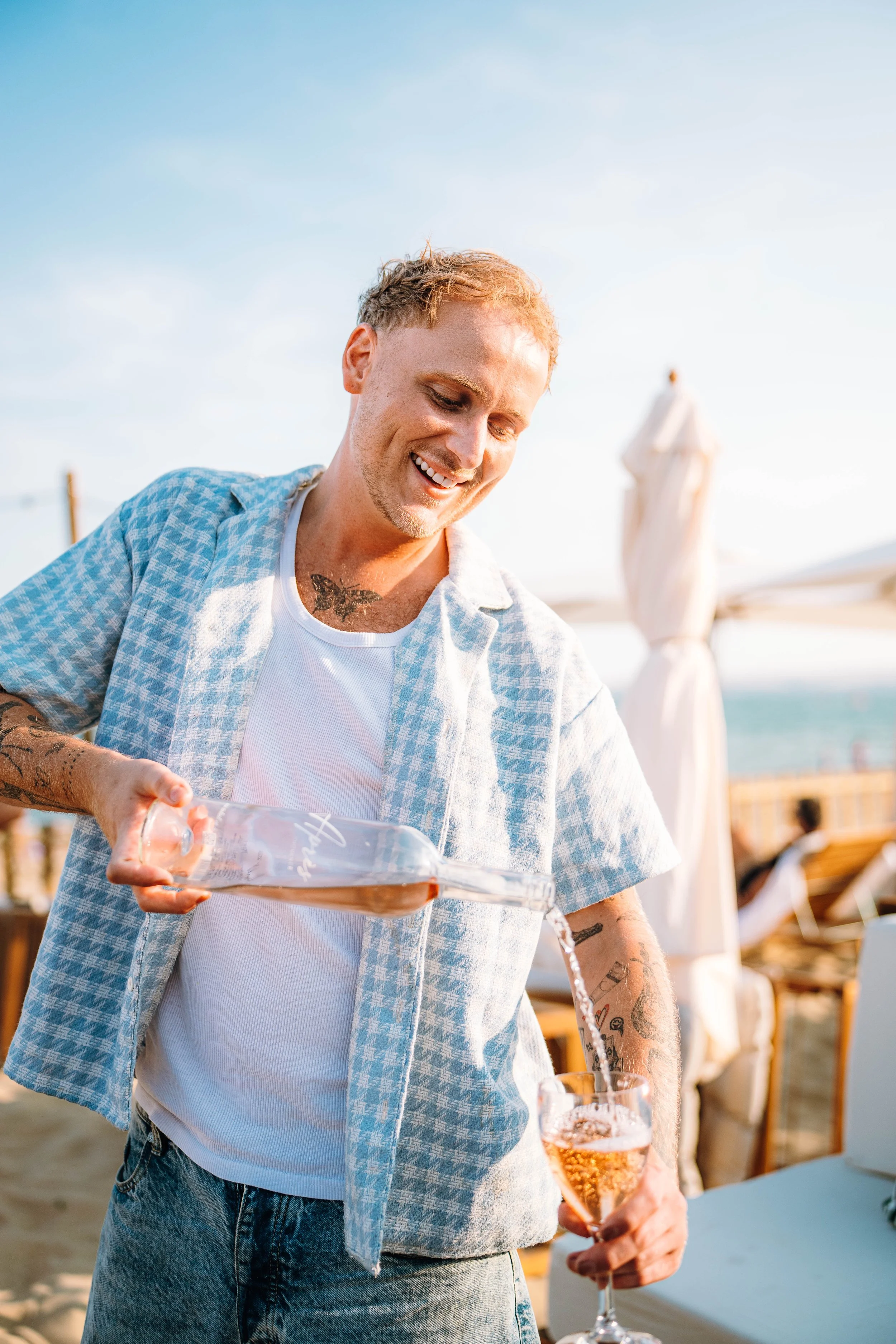 A young man with tattoos pouring water from a bottle into a glass at an outdoor event or beach, holding a glass of pink sparkling drink, wearing a casual shirt and jeans on a sunny day.