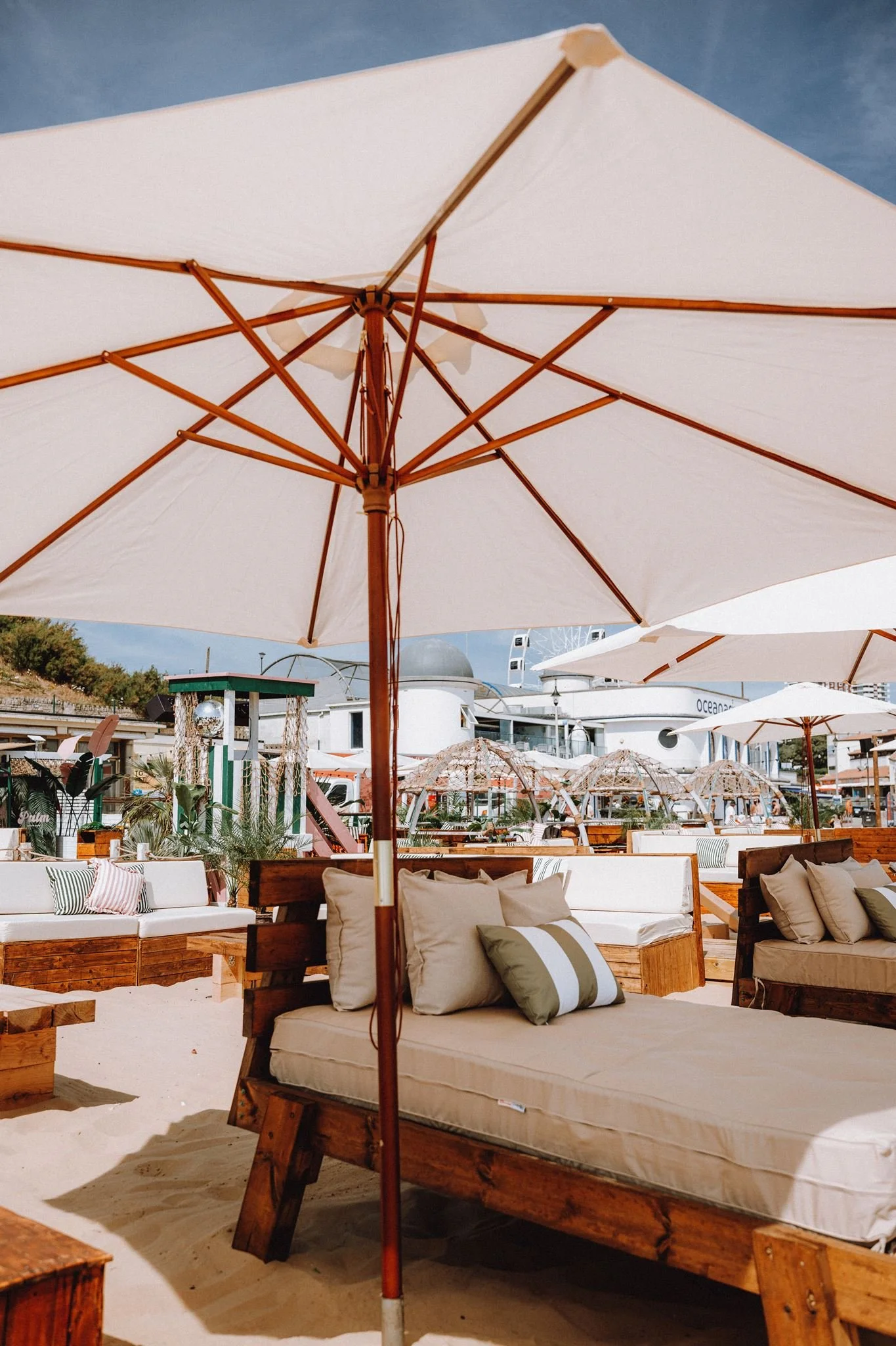 Beachside lounge area with large white umbrellas, wooden lounge chairs with cushions, and a sandy floor under a blue sky.