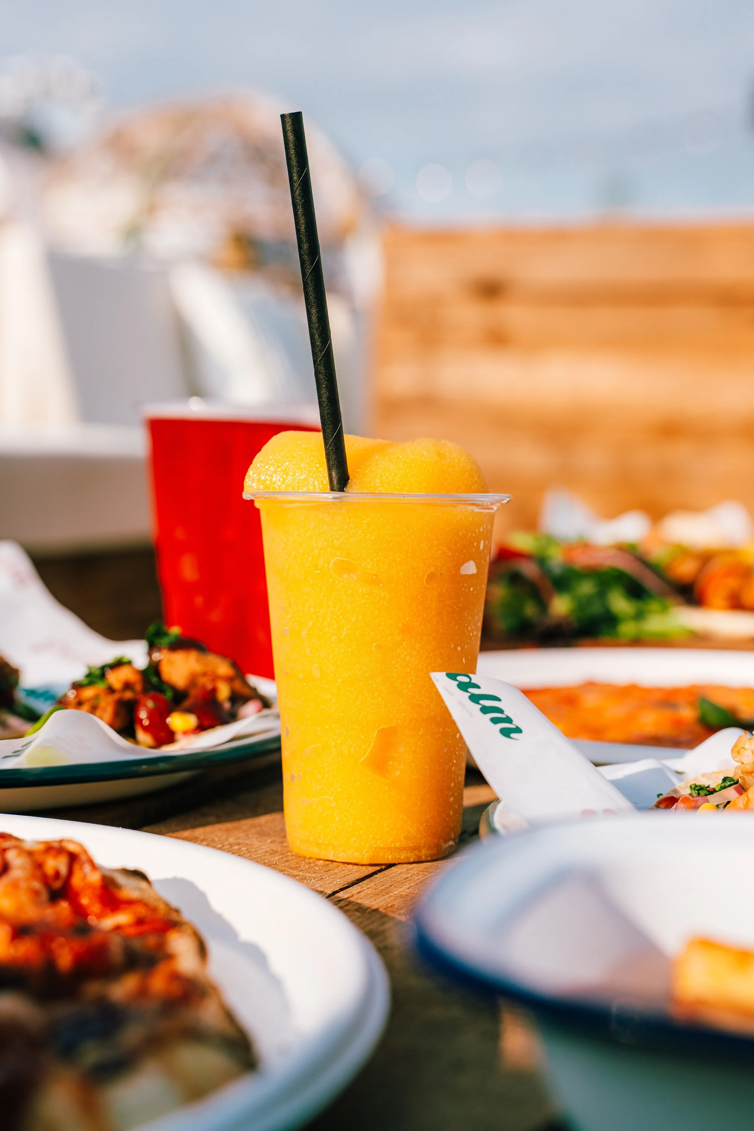 A yellow orange beverage with a black straw in a clear plastic cup placed on a wooden table, surrounded by plates of food at an outdoor meal setting with a blurred background including a body of water and sky.