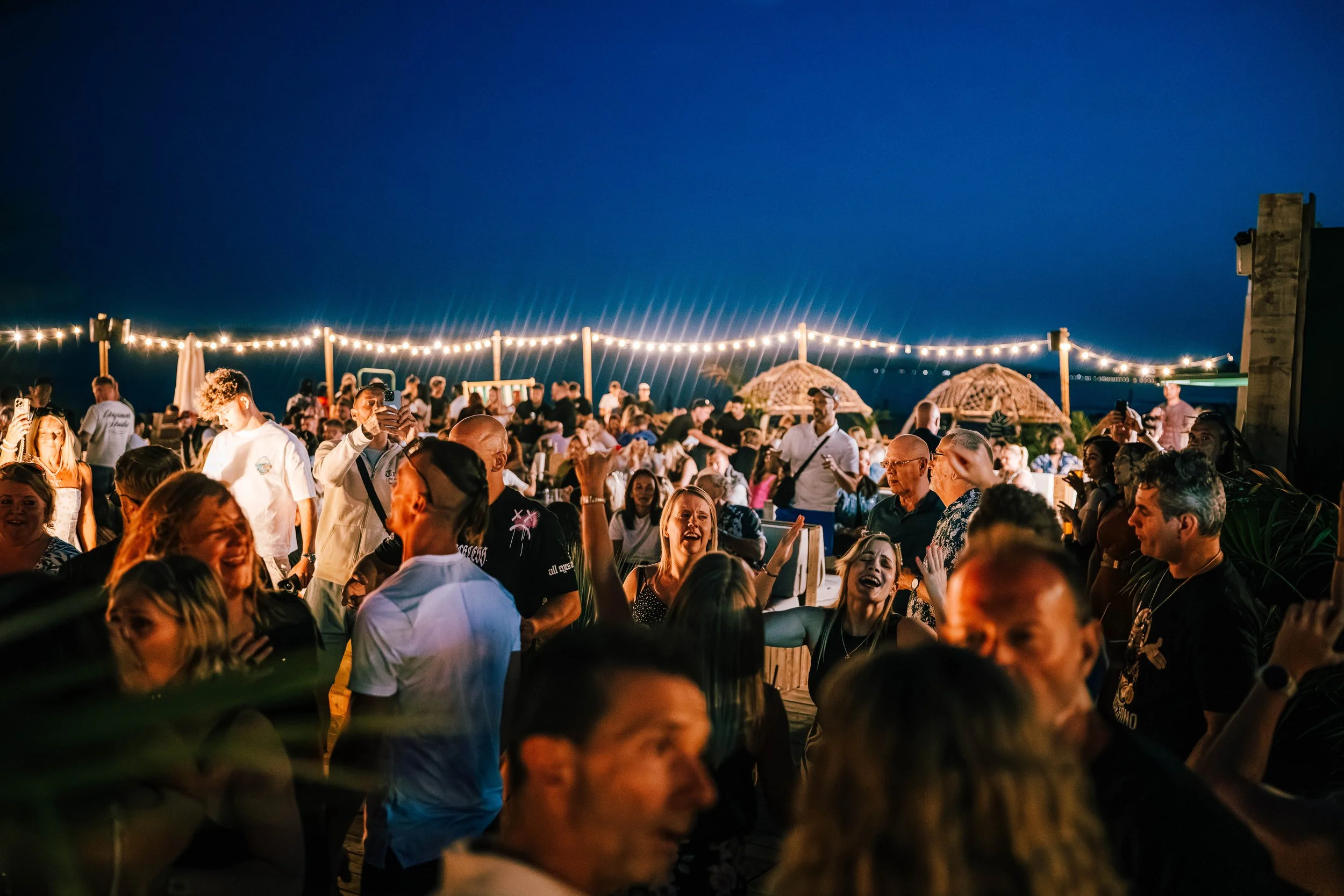 A large outdoor gathering with many people enjoying a night event under string lights, with a dark evening sky in the background.
