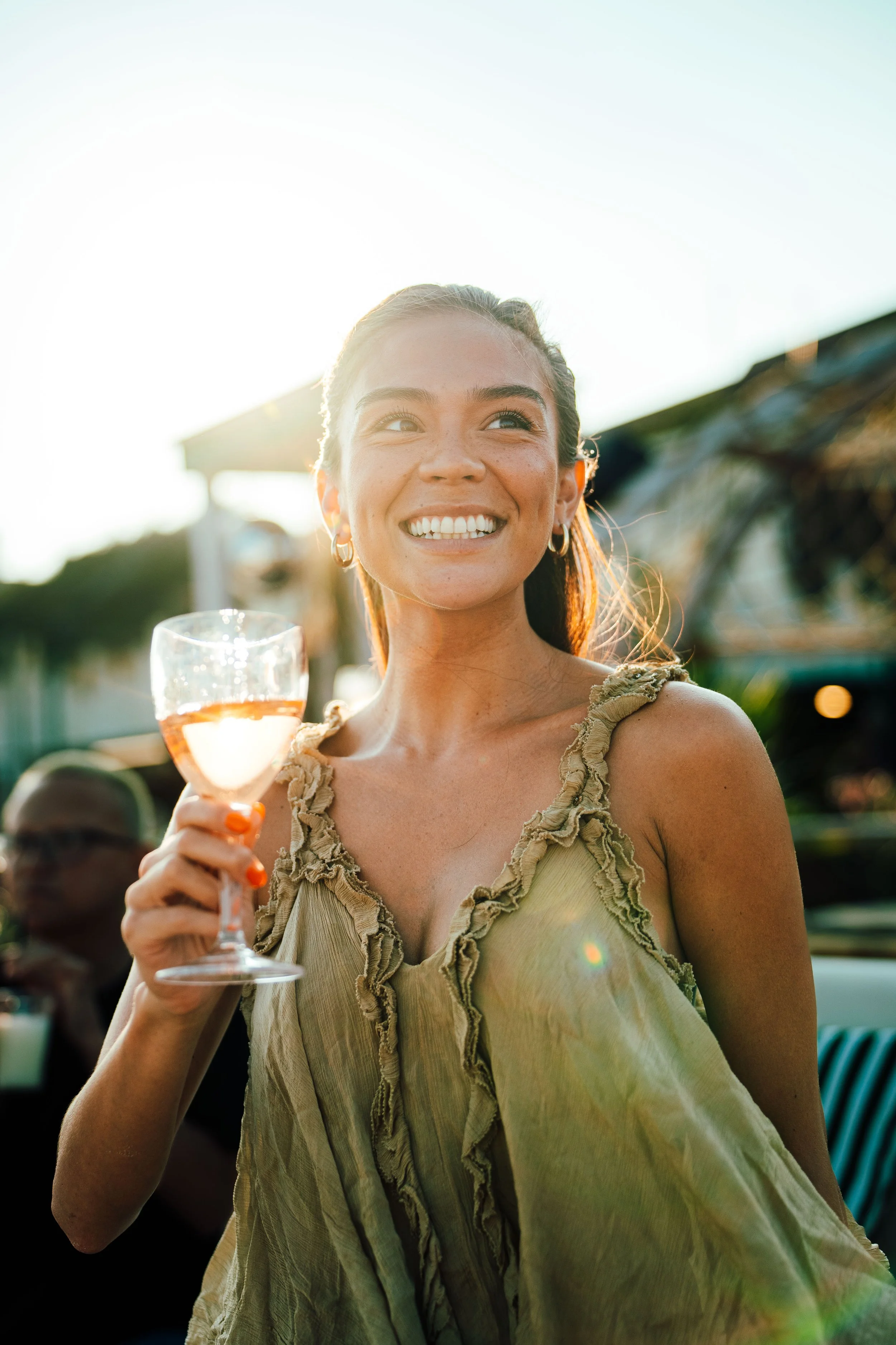 A woman with a big smile holding a glass of rose wine outdoors during daylight, wearing a beige ruffled top, with sunlight behind her and a blurred crowd in the background.