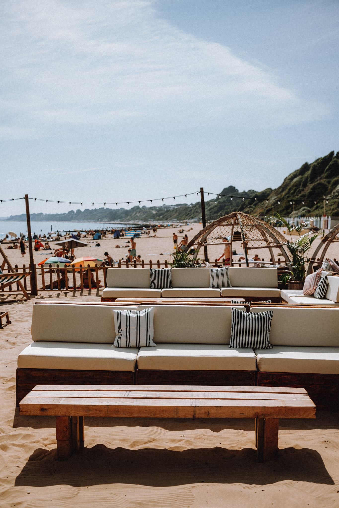 Beach scene with white sofas and striped cushions, wooden table, umbrellas, people, sand, ocean, and hillside in the background.