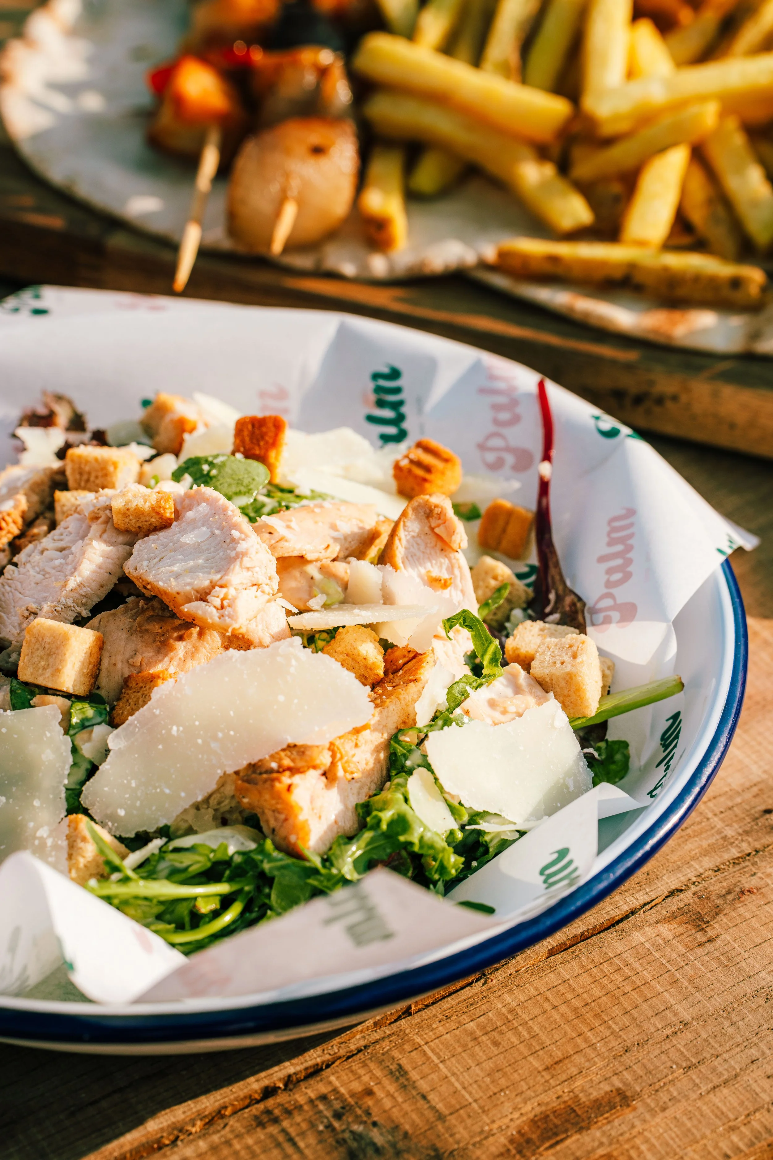 Caesar salad with lettuce, grilled chicken, croutons, and shaved cheese in a paper-lined bowl on a wooden table. In the background, there are French fries with skewered grilled sausages or meats.