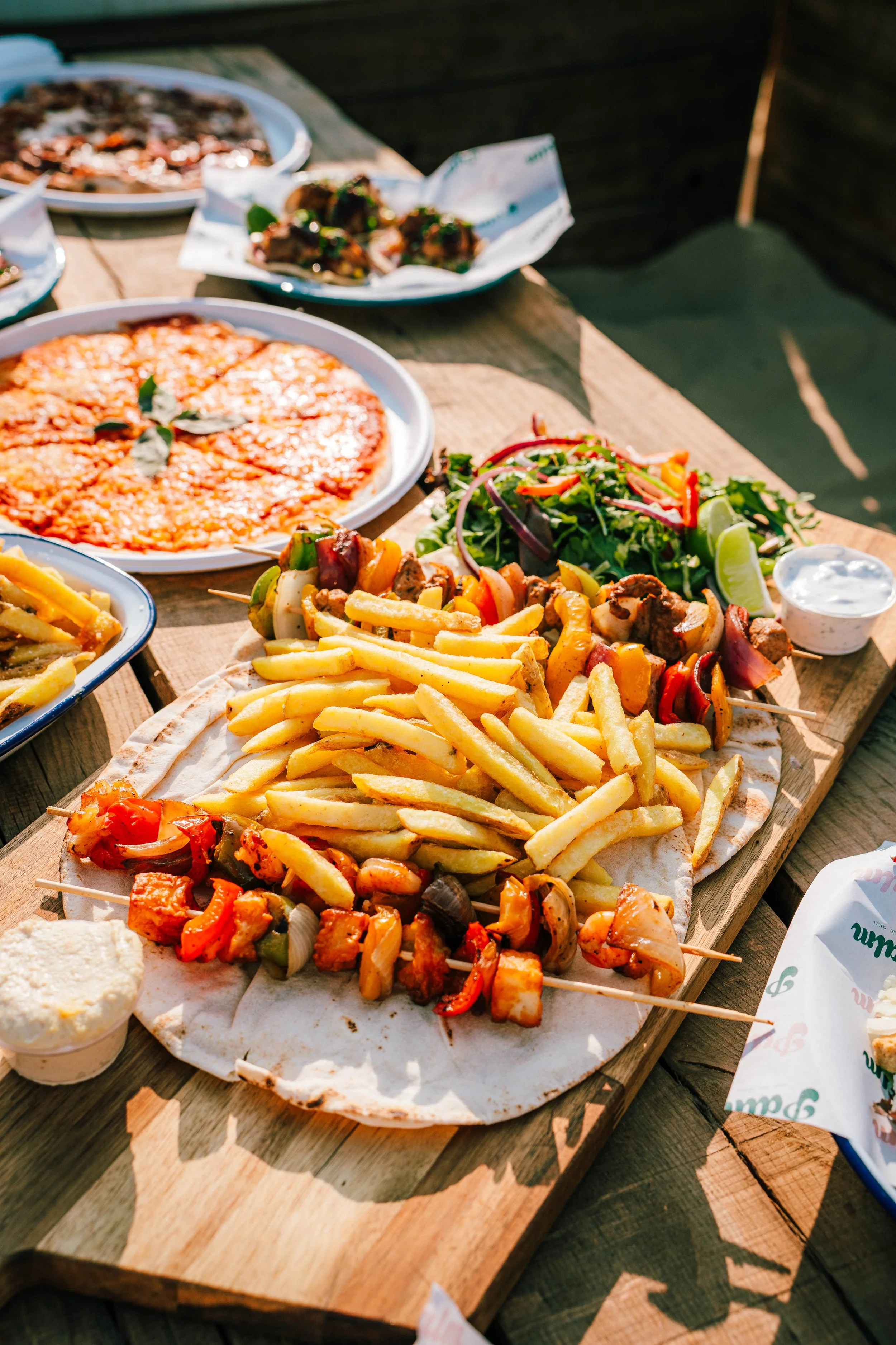 A wooden table with various dishes including French fries, grilled kebabs, salad with greens and vegetables, pizza, and additional plates of food in the background outdoors.