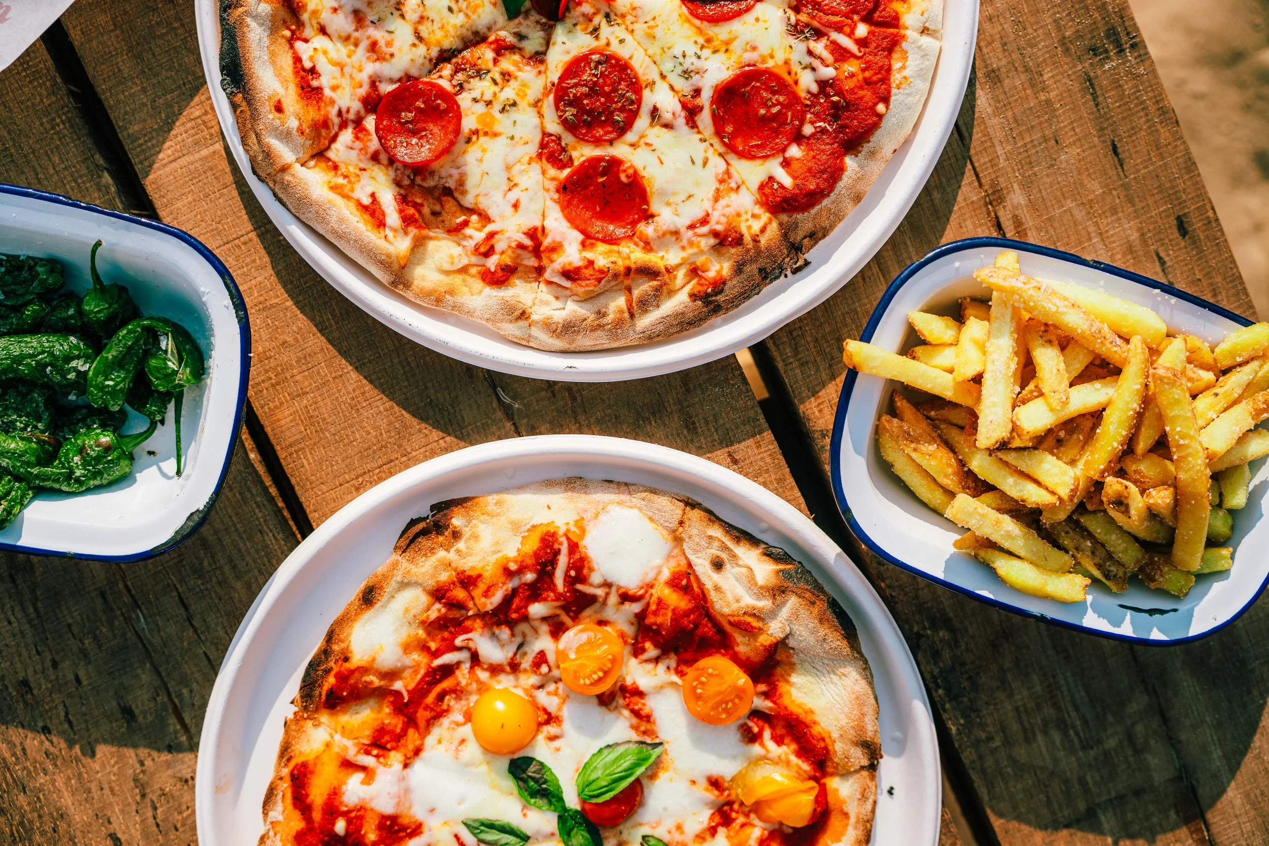 Two pizza slices with pepperoni and yellow tomatoes on a pizza with cheese, and a pizza slice with eggs, cheese, tomato, and basil. Also, a bowl of French fries and a bowl of roasted green peppers on a wooden table.