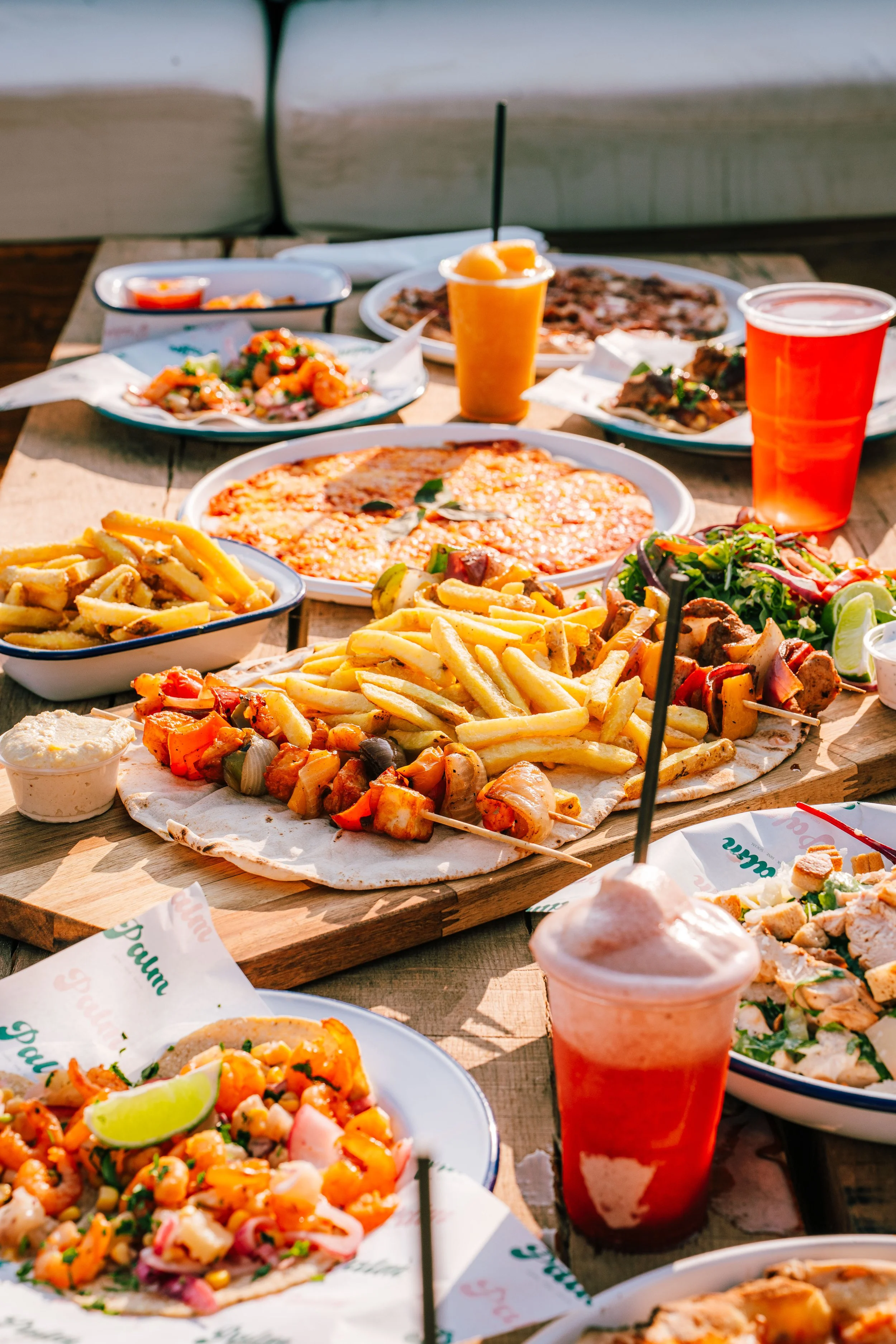 A table of various Mexican dishes including pizza, tacos, nachos, a salad, and beverages such as smoothies and drinks, on a wooden table outdoors.
