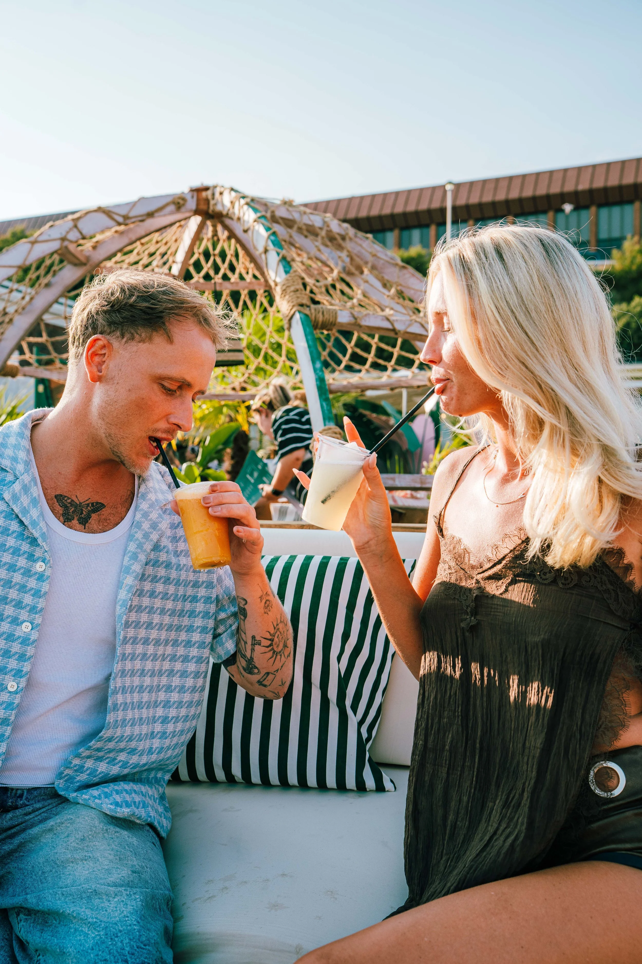 A man and woman sitting outdoors, both sipping drinks through straws and enjoying each other's company. The man has tattoos on his arm and chest, wearing a checkered shirt, while the woman has long blonde hair and is wearing a dark lace dress. In the background, there is a woven umbrella, greenery, and a building.
