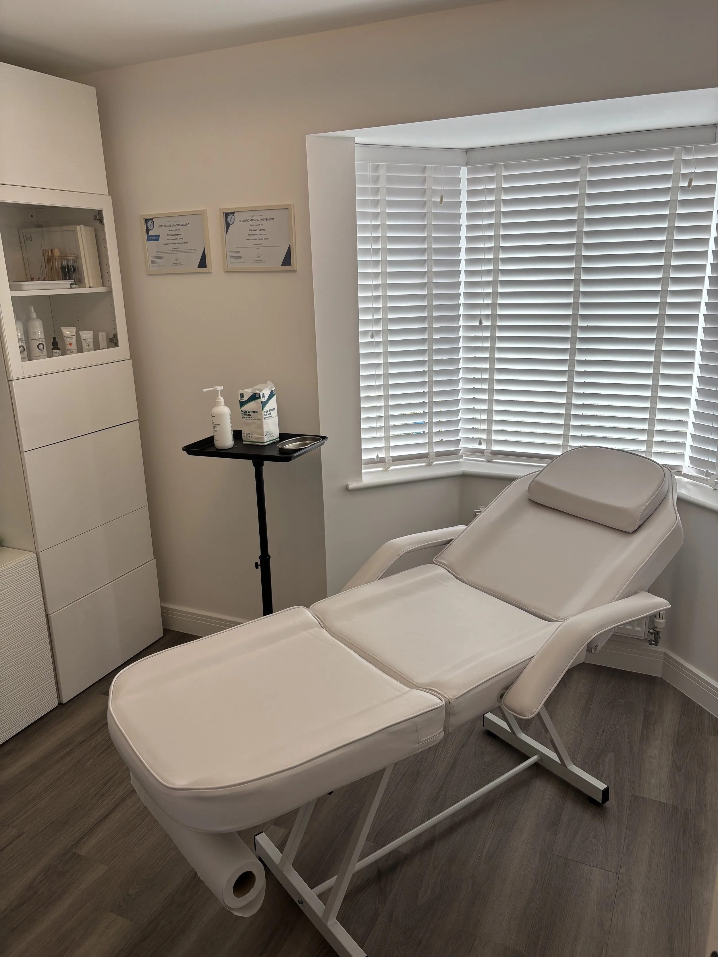 Medical examination room with a white adjustable reclining chair, medicine cabinet, and window with white blinds.