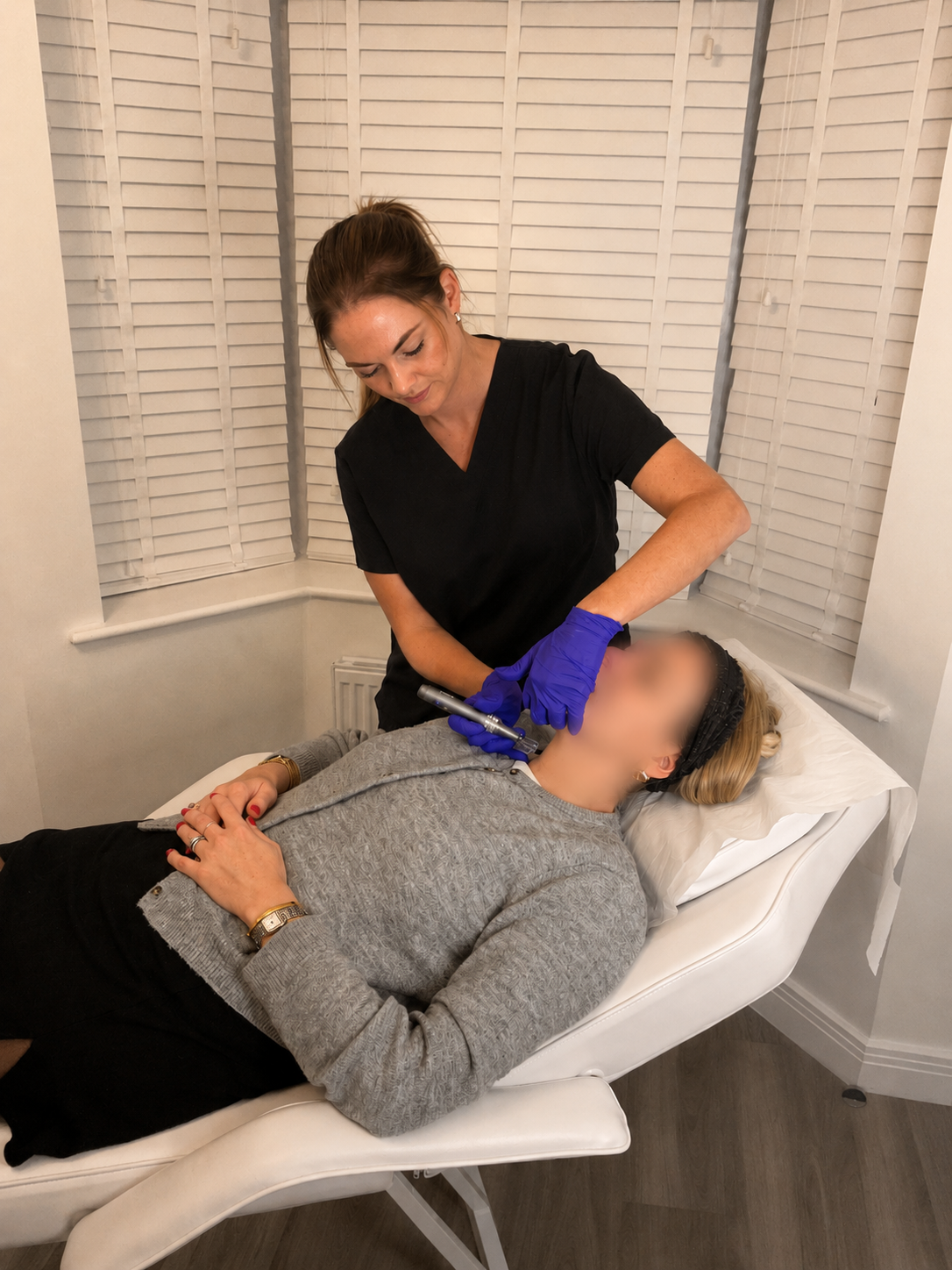 A woman lying on a medical treatment bed being administered a facial treatment by a female esthetician. The esthetician is wearing black scrubs and purple gloves, and the woman is dressed in a grey sweater and black pants, with her hands resting on her stomach.