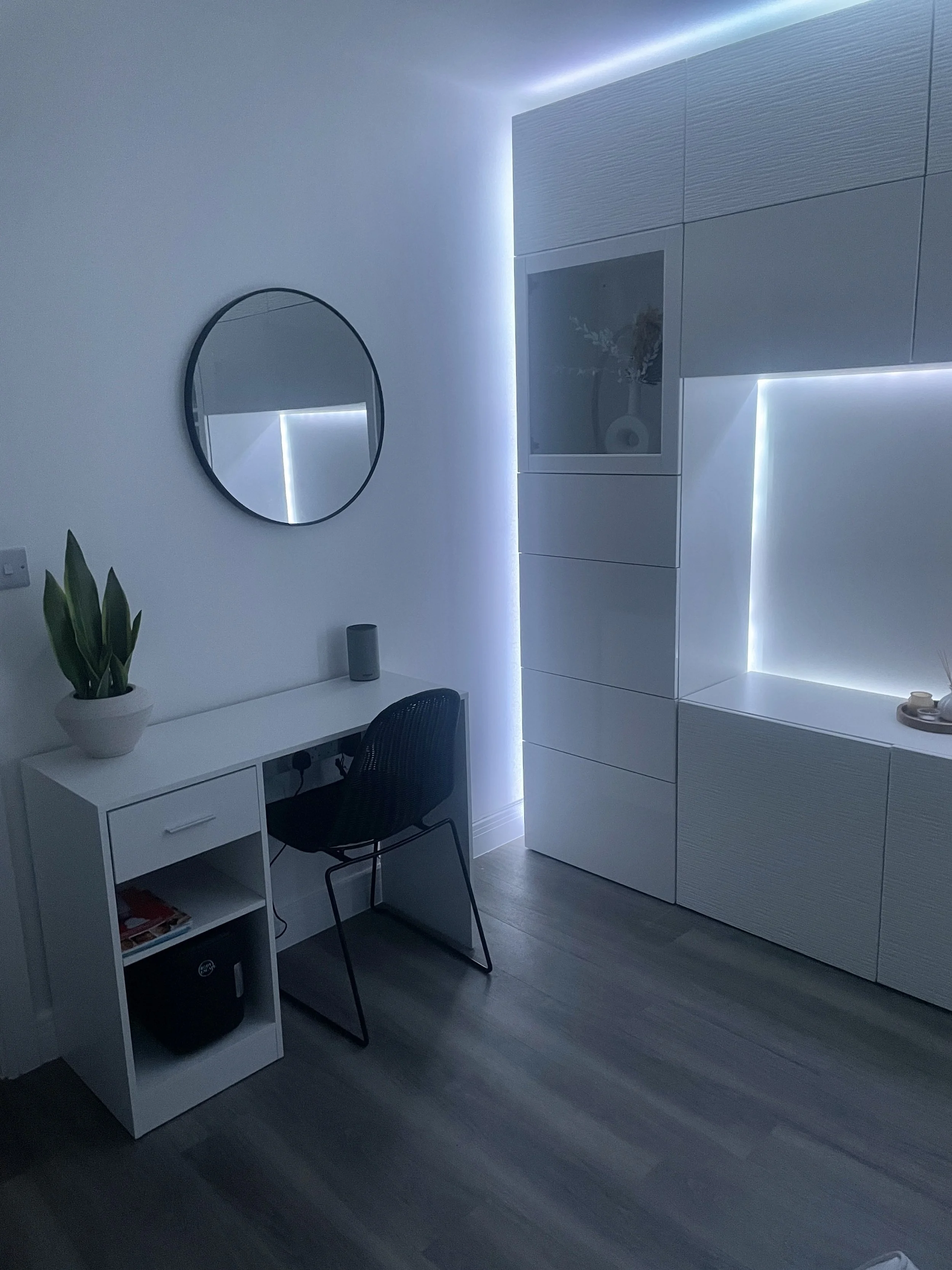 A minimalist modern room featuring a white desk with a potted plant, a black chair, a mirror on the wall, and white cabinets with LED lighting accents.