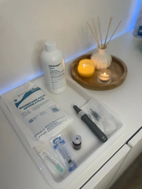 Medical and skincare items on a white surface, including a bottle of skin cleanser, a pack of wound-care dressings, a black skincare tool, a small dropper bottle, and a tube of ointment, with a decorative tray holding a candle, a candle holder, and reed diffuser sticks in the background.
