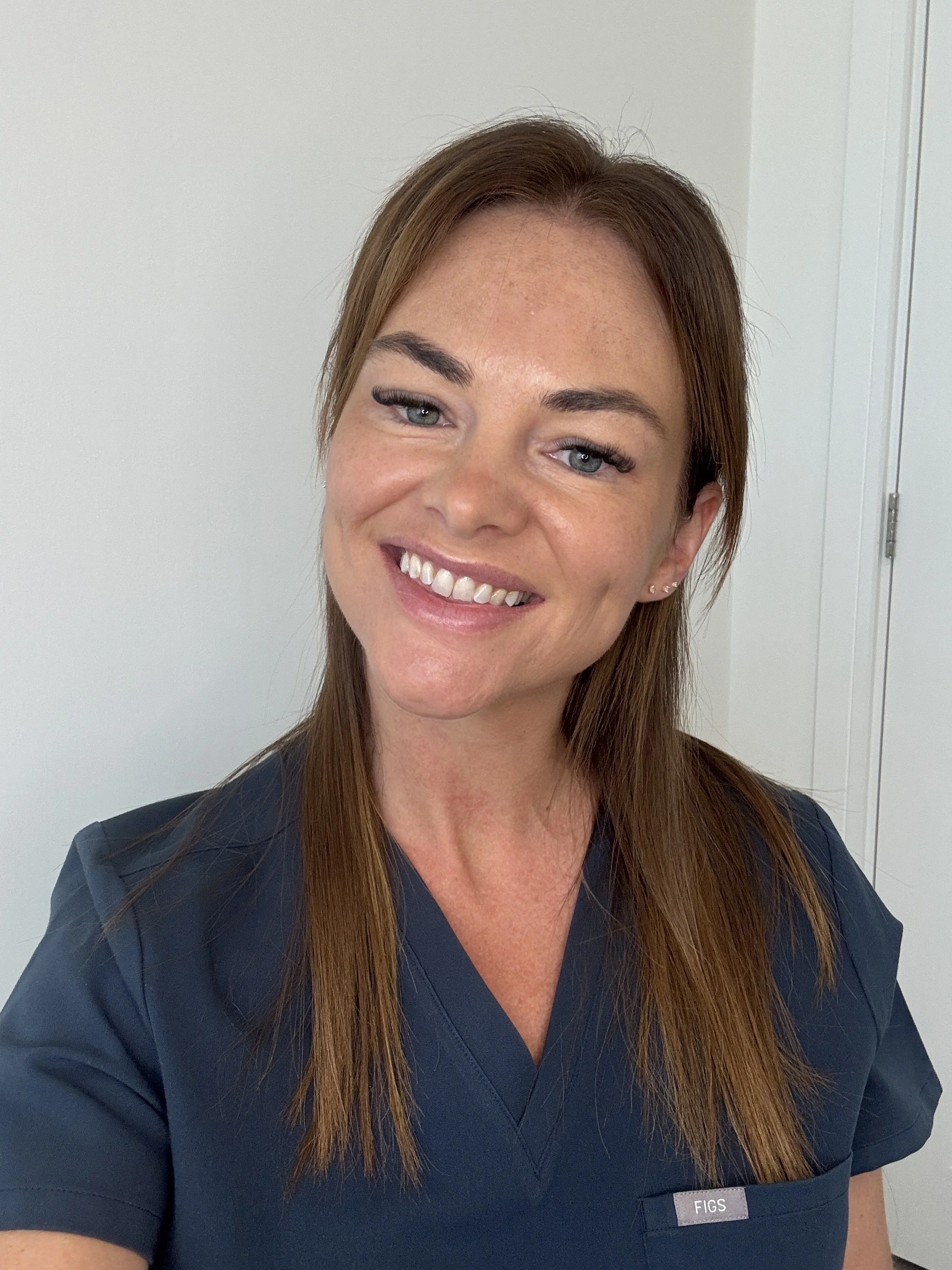 Close-up of a smiling woman with long brown hair wearing a dark blue medical uniform with a name tag that reads 'FIGS', standing against a plain light-colored wall.