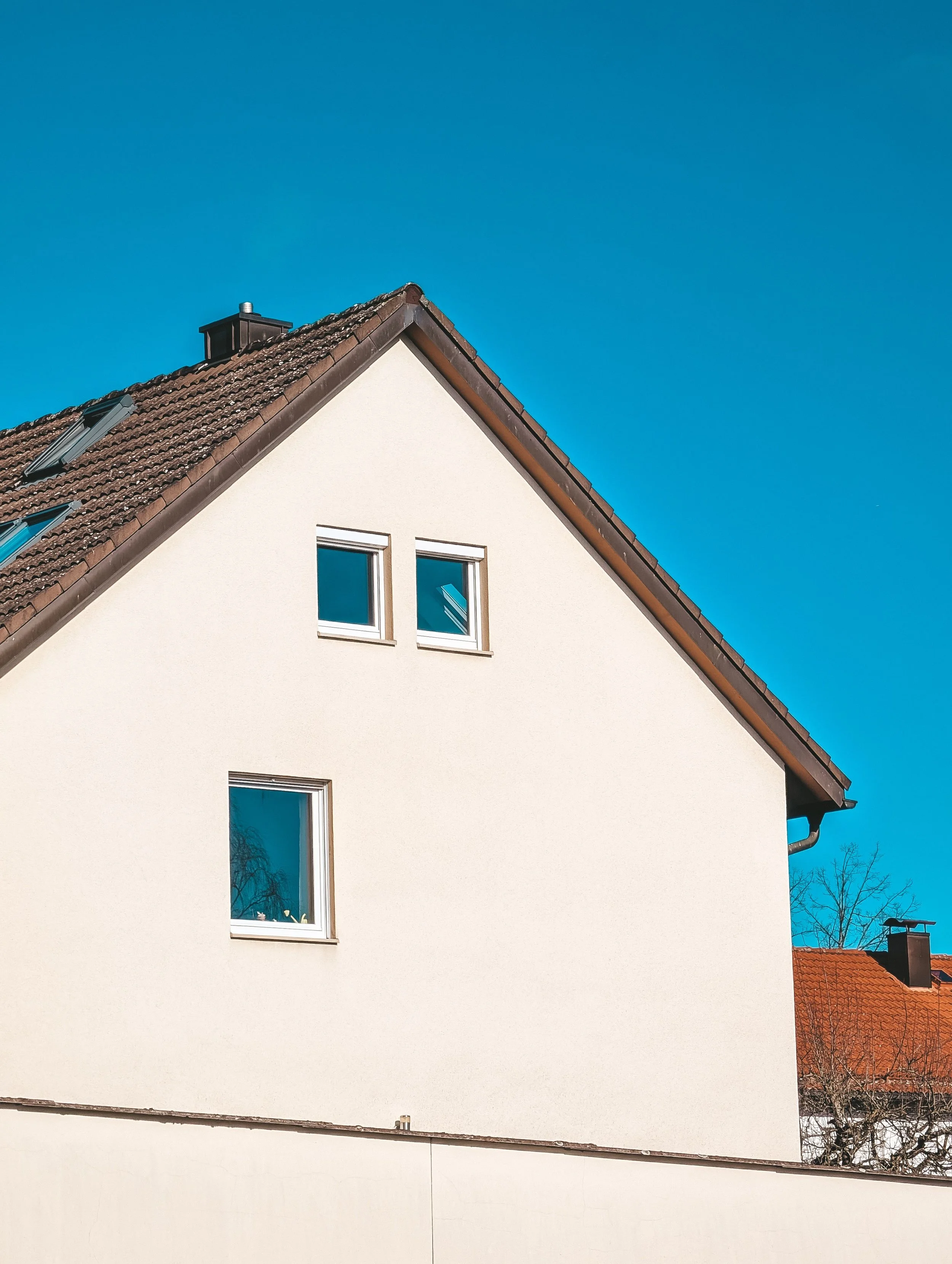 The upper part of a white residential building with a sloped brown tiled roof, two small rectangular windows, and a chimney, under a clear blue sky.