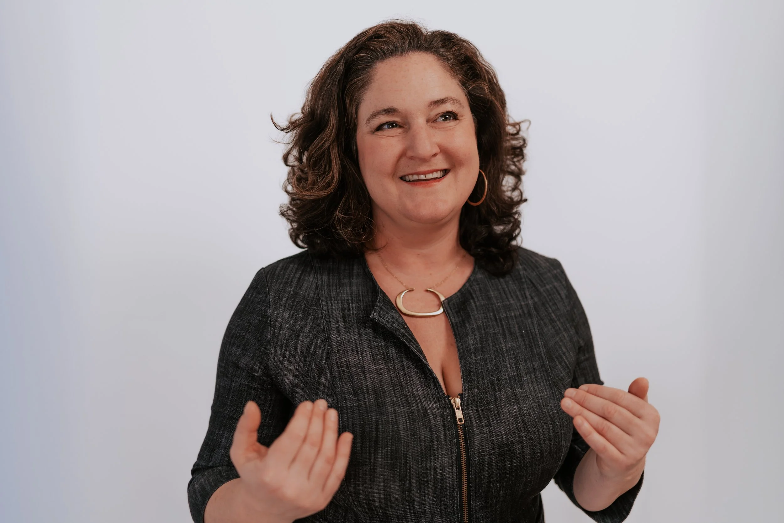 A woman with curly brown hair, wearing a black zip-up jacket, is smiling and gesturing with her hands against a plain white background.