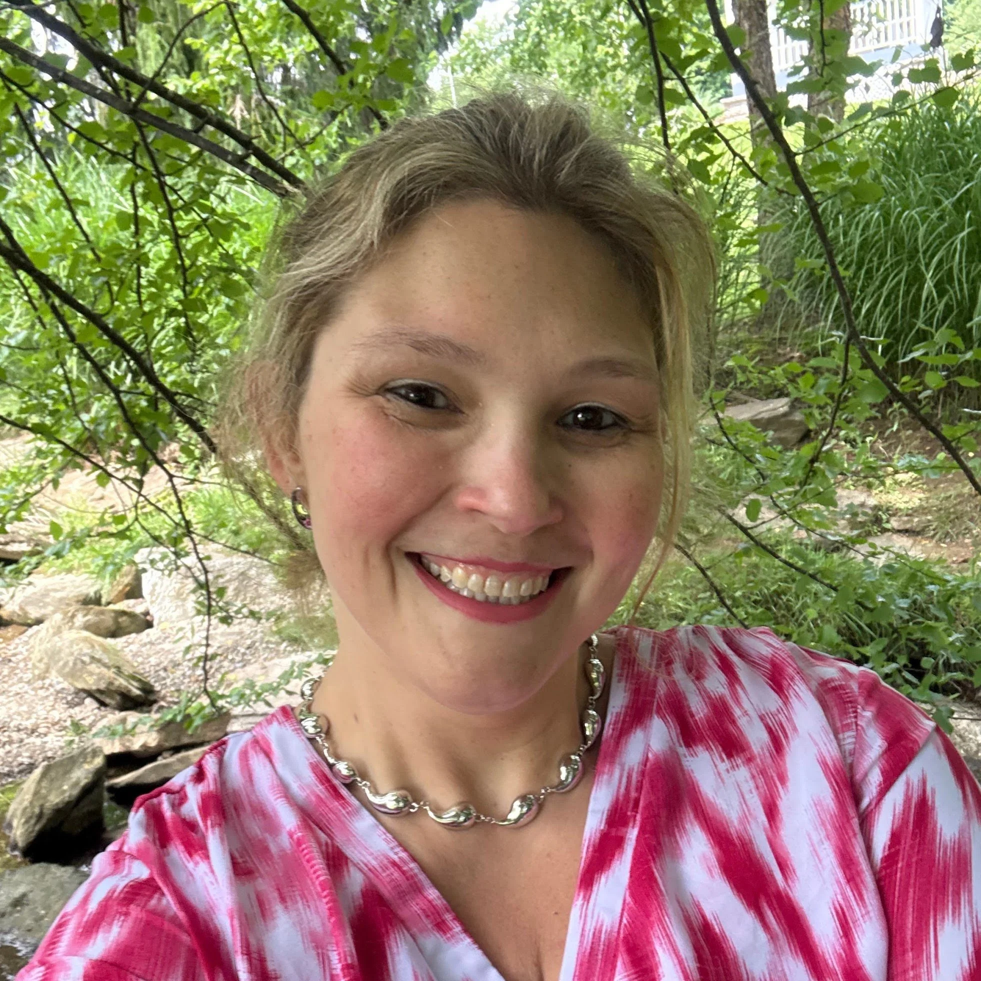 Close-up of a woman's face, showing her smile, eyes, and light brown hair with blonde highlights, wearing a pink and white patterned top and a silver necklace, outdoors with a green wooded background.