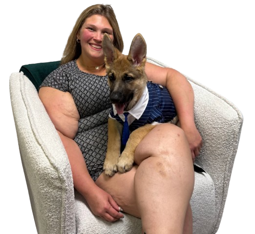 A woman with long blonde hair sitting on a white armchair, smiling, holding a German Shepherd puppy wearing a blue and white striped shirt and a tie.