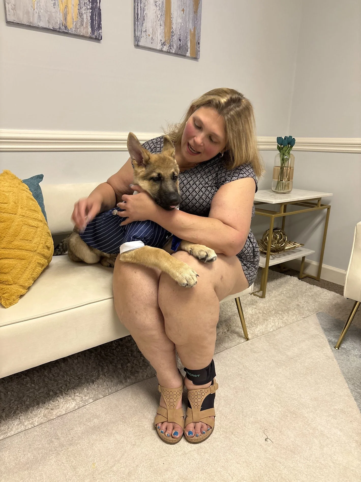 A woman sitting on a white couch holding a puppy, which is wearing a striped shirt. The woman has blond hair, and is wearing a patterned dress, tan high-heeled shoes, and a knee brace.