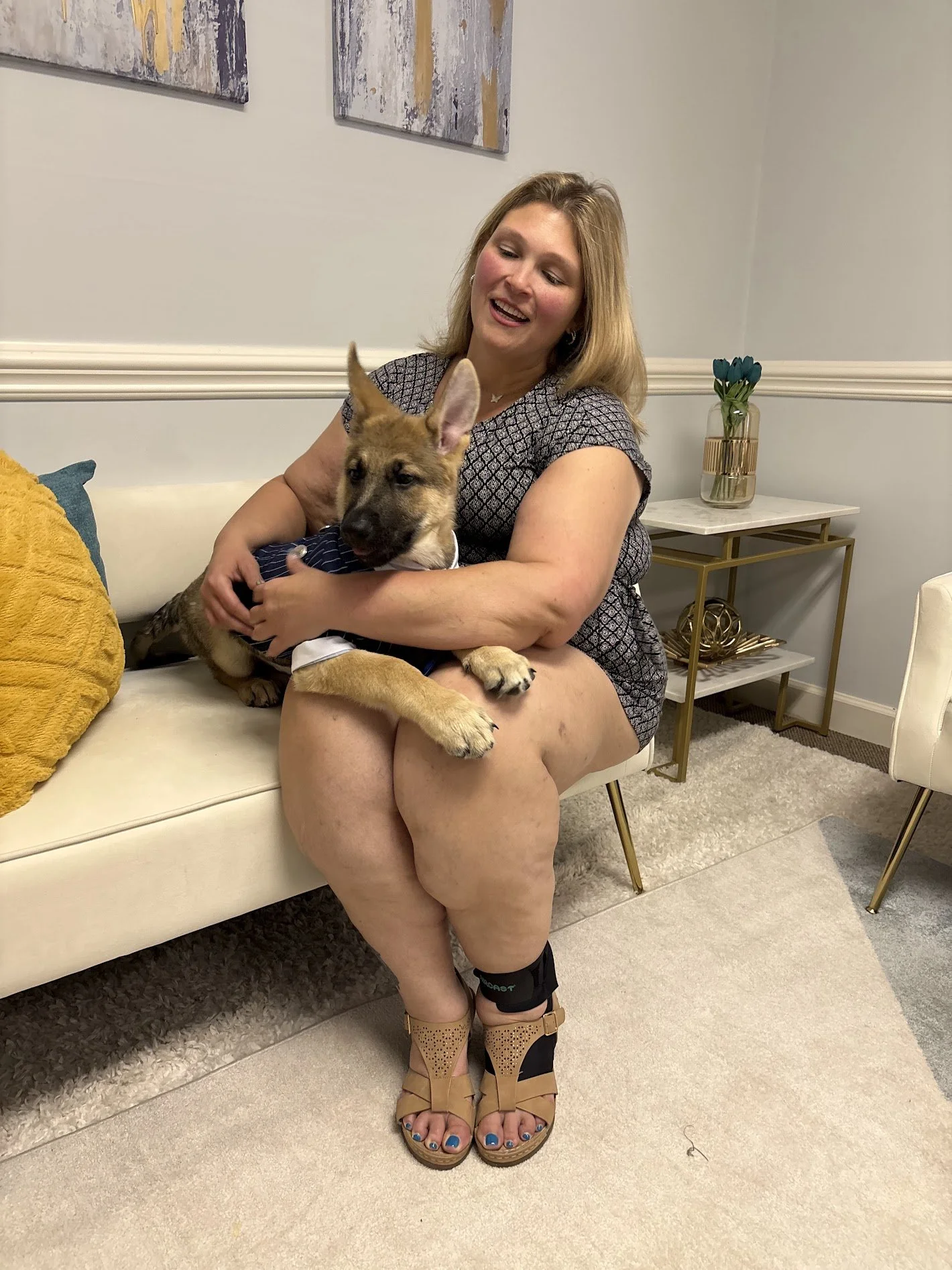 A woman is sitting on a white couch holding a German Shepherd puppy wearing a striped shirt. She is smiling and wearing a patterned dress, tan sandals, and has a knee brace on her left leg. There is a yellow pillow on the couch, and a side table with a vase of blue flowers behind her. The room has light-colored walls with abstract artwork hanging on the wall.