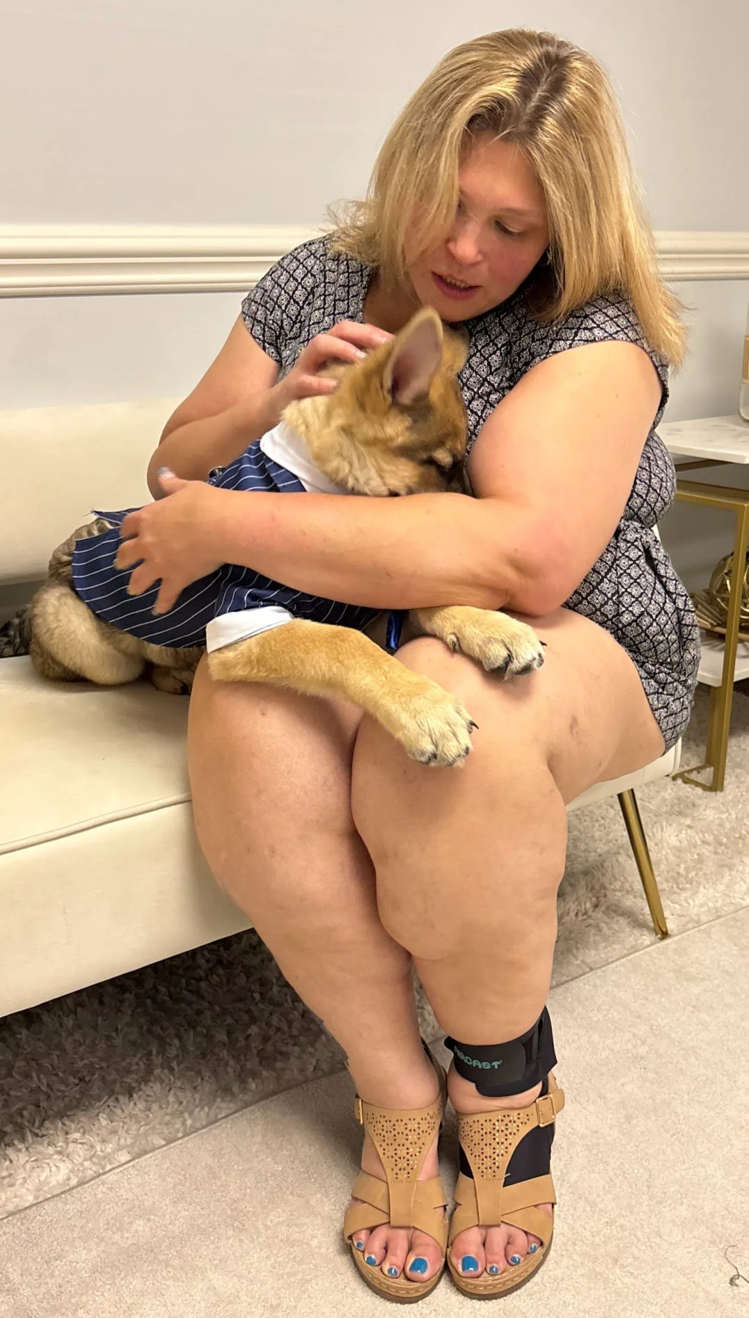A woman sitting on a beige sofa holding a puppy dressed in a blue striped outfit on her lap. The woman is wearing a patterned dress and tan wedge sandals, with her legs crossed. The puppy looks calm, and the woman appears to be gently petting or cuddling it.
