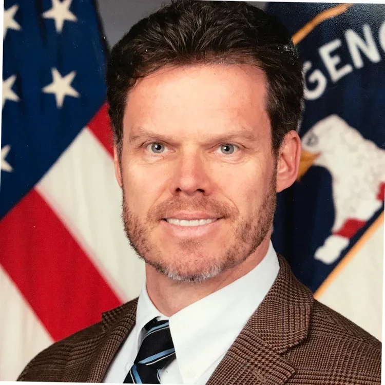 A man wearing a blue and white checkered shirt with a pink tie, standing against a plain gray background.