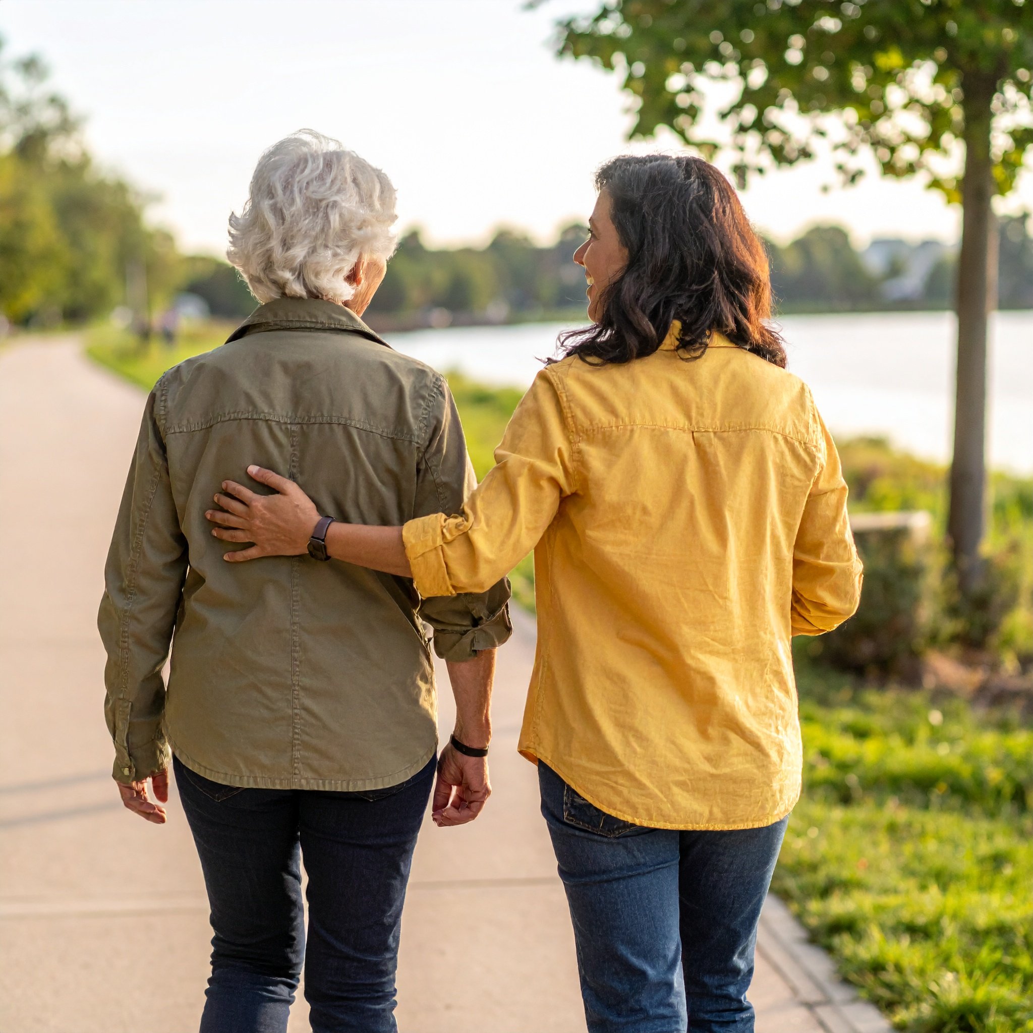 Two women walking side by side outdoors, one with curly white hair wearing a khaki jacket, and the other with dark curly hair wearing a yellow jacket, holding each other around the shoulders.